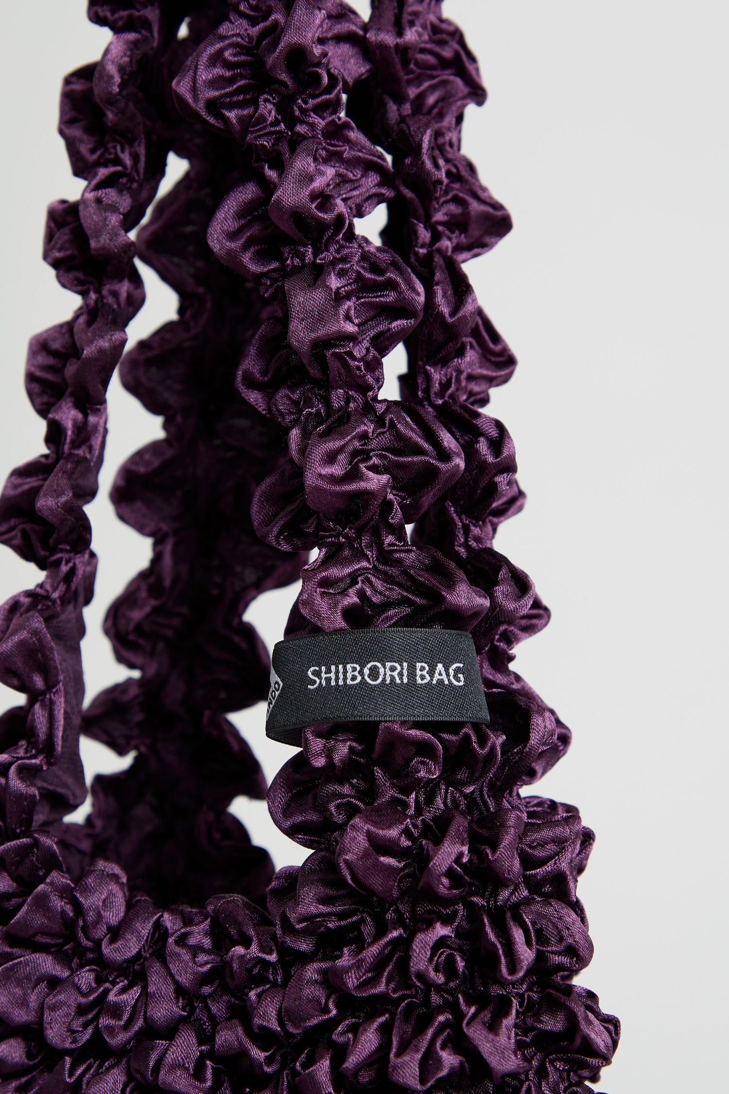 Close-up of an Aubergine Shibori Pleated Bag, featuring a deep purple, gathered handle and a small black label that reads “SHIBORI BAG”.