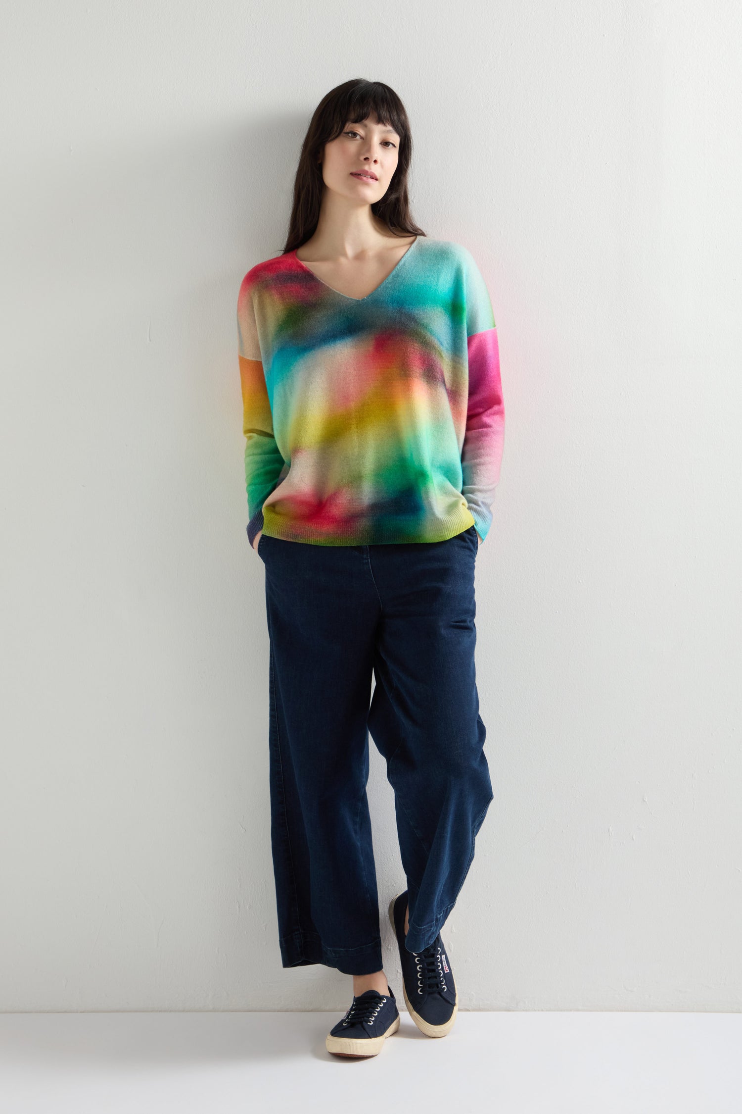 Against a plain white background, someone gracefully dons a V-Neck Cashmere Jumper with a lively rainbow watercolour print paired with dark pants.