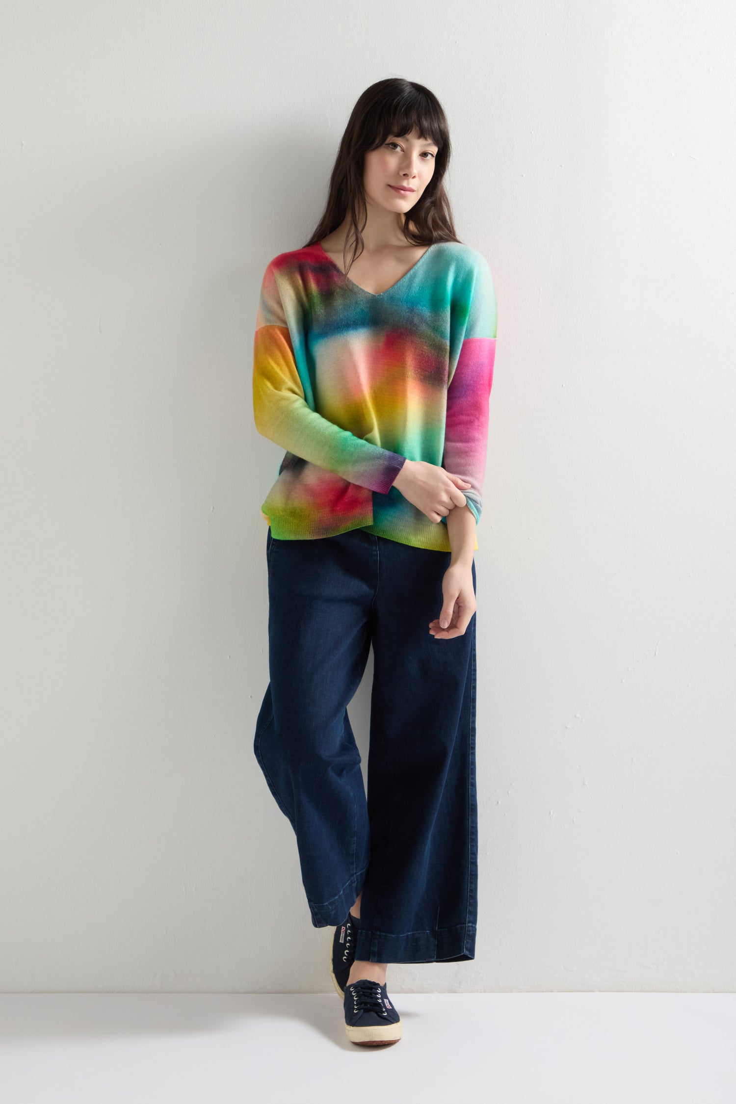 A person stands against a white wall, wearing a V-Neck Cashmere Jumper with a drop shoulder design and dark wide-leg pants, one hand resting on their other arm.