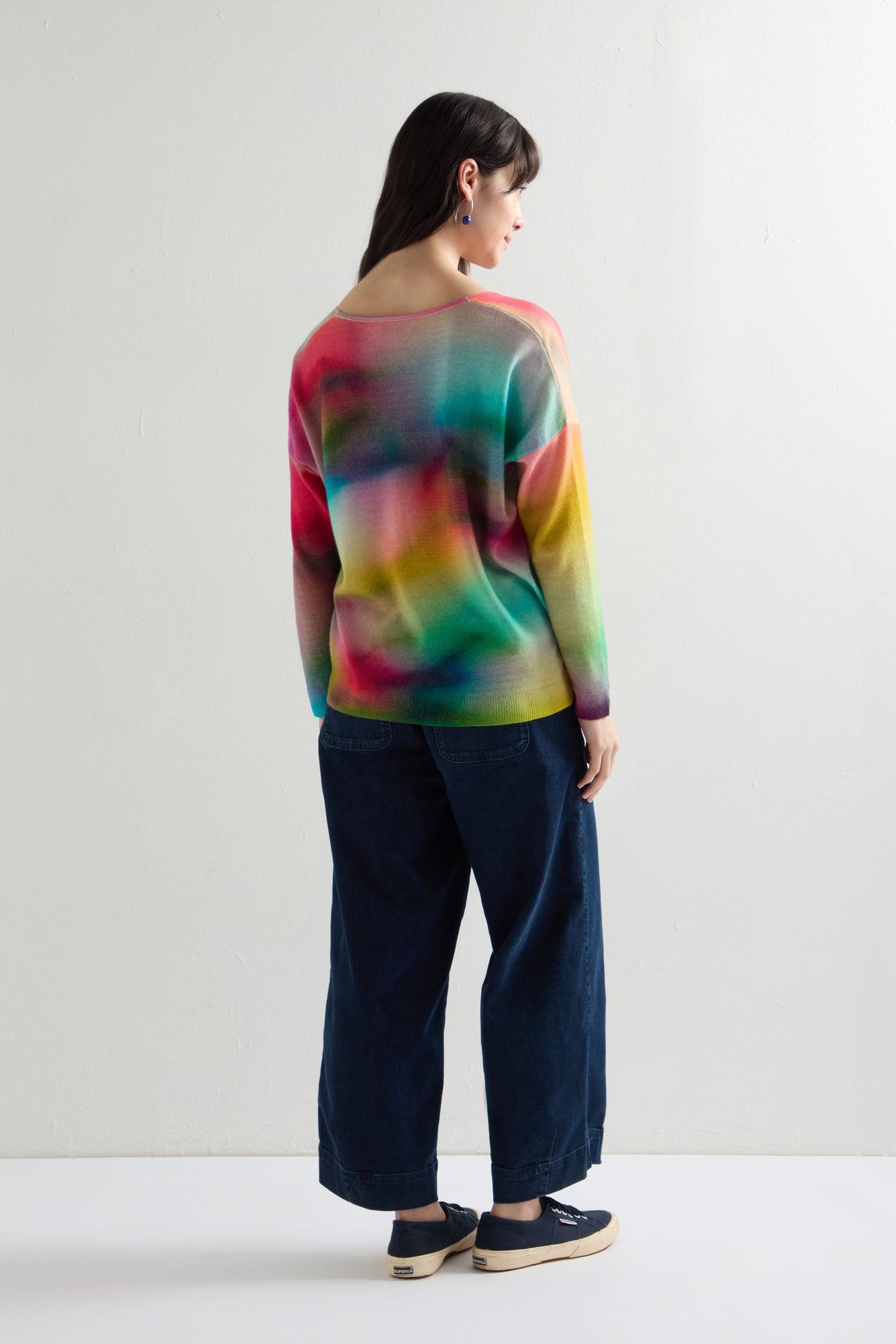 A person in a vibrant rainbow V-Neck Cashmere Jumper with drop shoulders, dark wide-leg jeans, and navy sneakers stands against a plain white background.