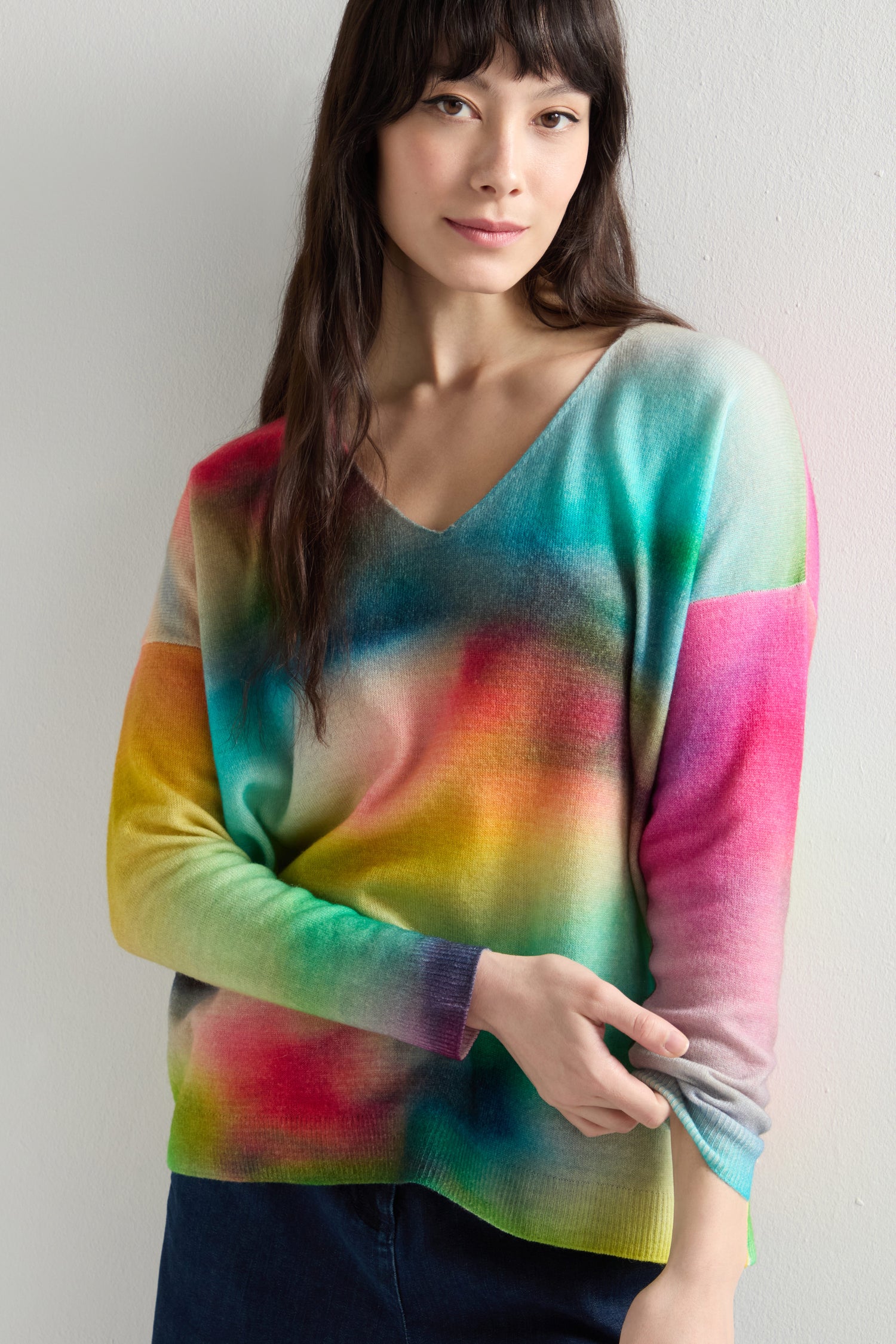 A person stands against a plain background, wearing a vibrant V-Neck Cashmere Jumper featuring a gradient design, drop shoulders, and long sleeves. The rainbow watercolor print enhances the artistic flair of their outfit.