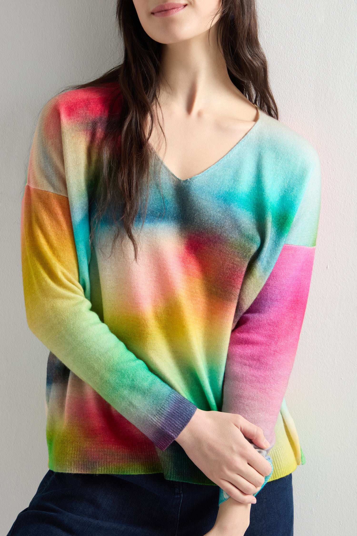 Against a neutral background, a person wears the V-Neck Cashmere Jumper featuring a vibrant rainbow watercolor print and drop shoulder design.