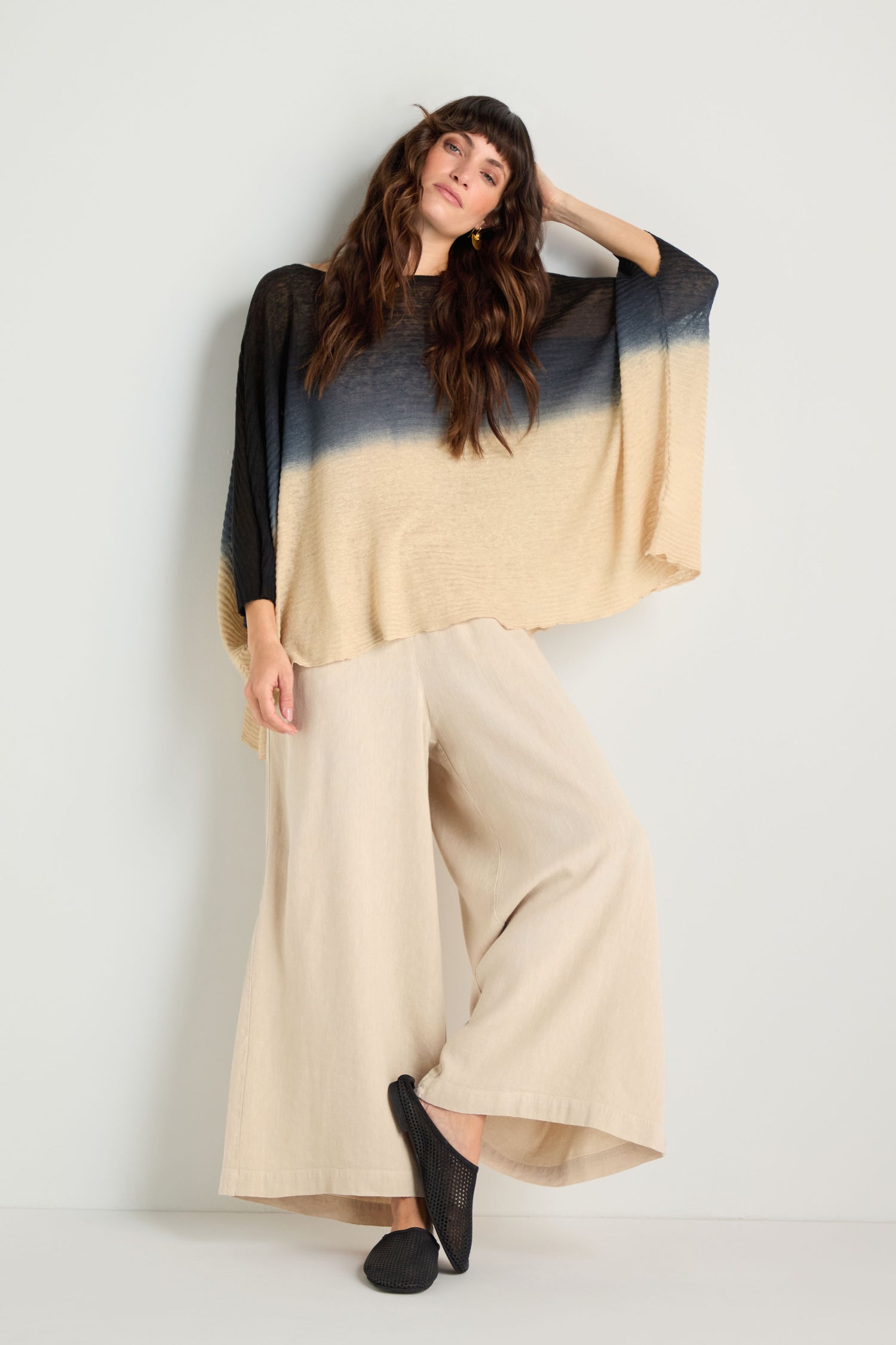 A woman stands against a white wall wearing the Dip Dye Knit Easy Top, a loose black-to-beige linen knit, paired with wide beige pants and black slip-on shoes.
