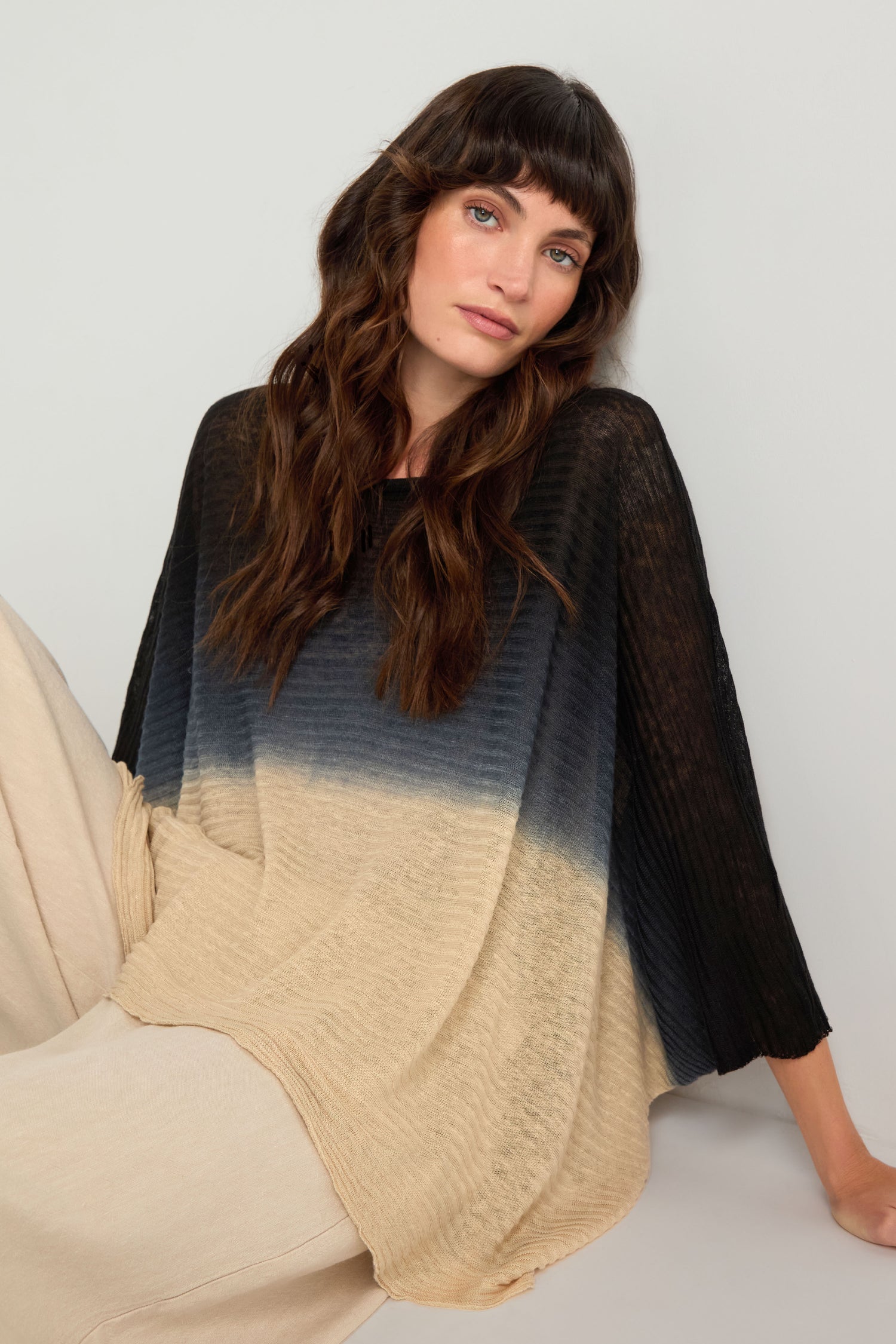 A woman with long brown hair wears the Dip Dye Knit Easy Top in black and beige with light pants, sitting against a light background.