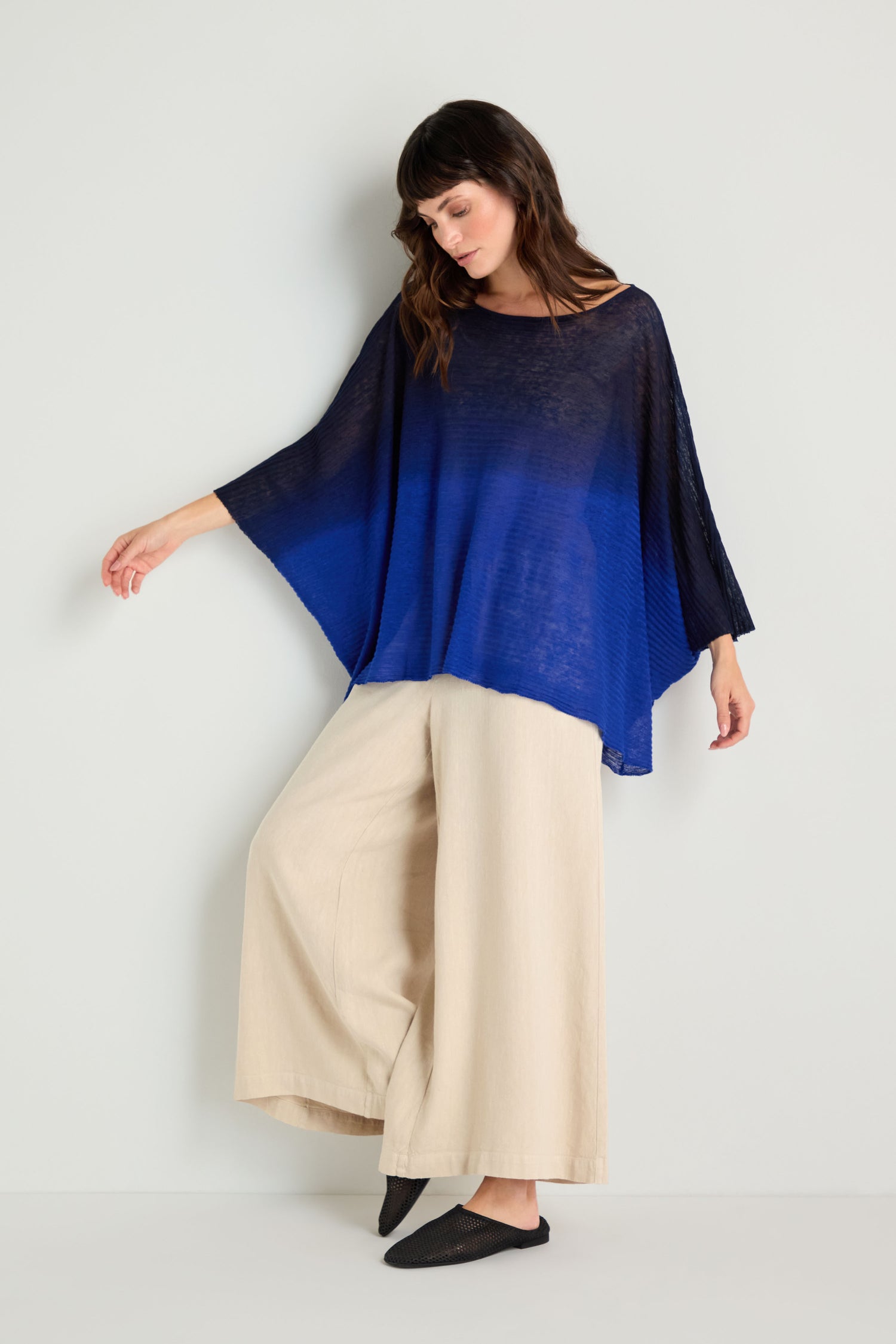 A woman stands against a white wall wearing the Navy Blue Dip Dye Knit Easy Top, wide-legged beige pants, and black slip-on shoes for a relaxed silhouette.