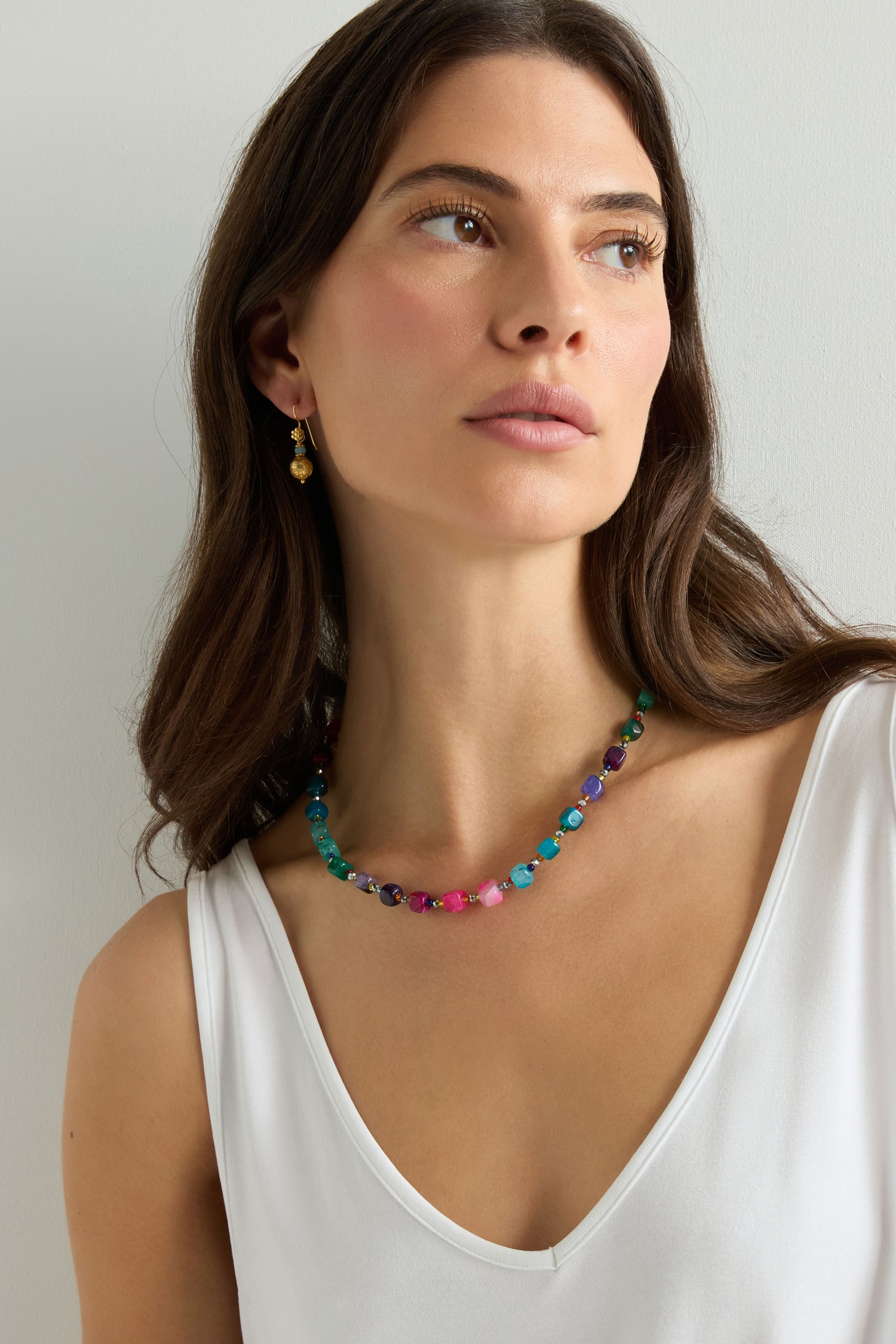 A woman in a sleeveless white top and gold earrings wears the Artisanal Rainbow Agate Cube Necklace, looking slightly to the side against a plain background.