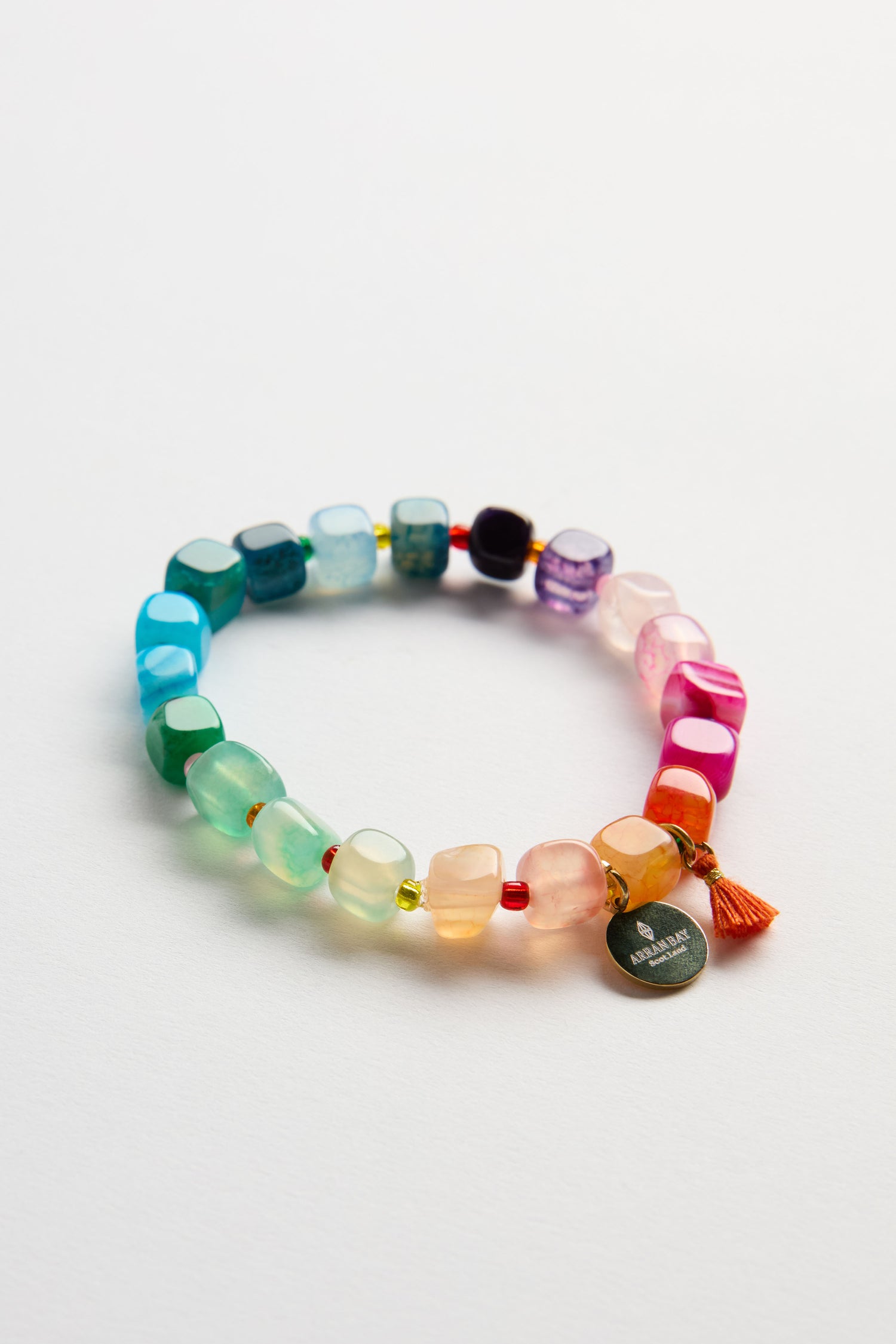 The Artisanal Rainbow Agate Bead Bracelet features multicolored translucent agate beads, a small orange tassel, and a round metallic tag, displayed on a white background—a vibrant handcrafted jewelry piece.