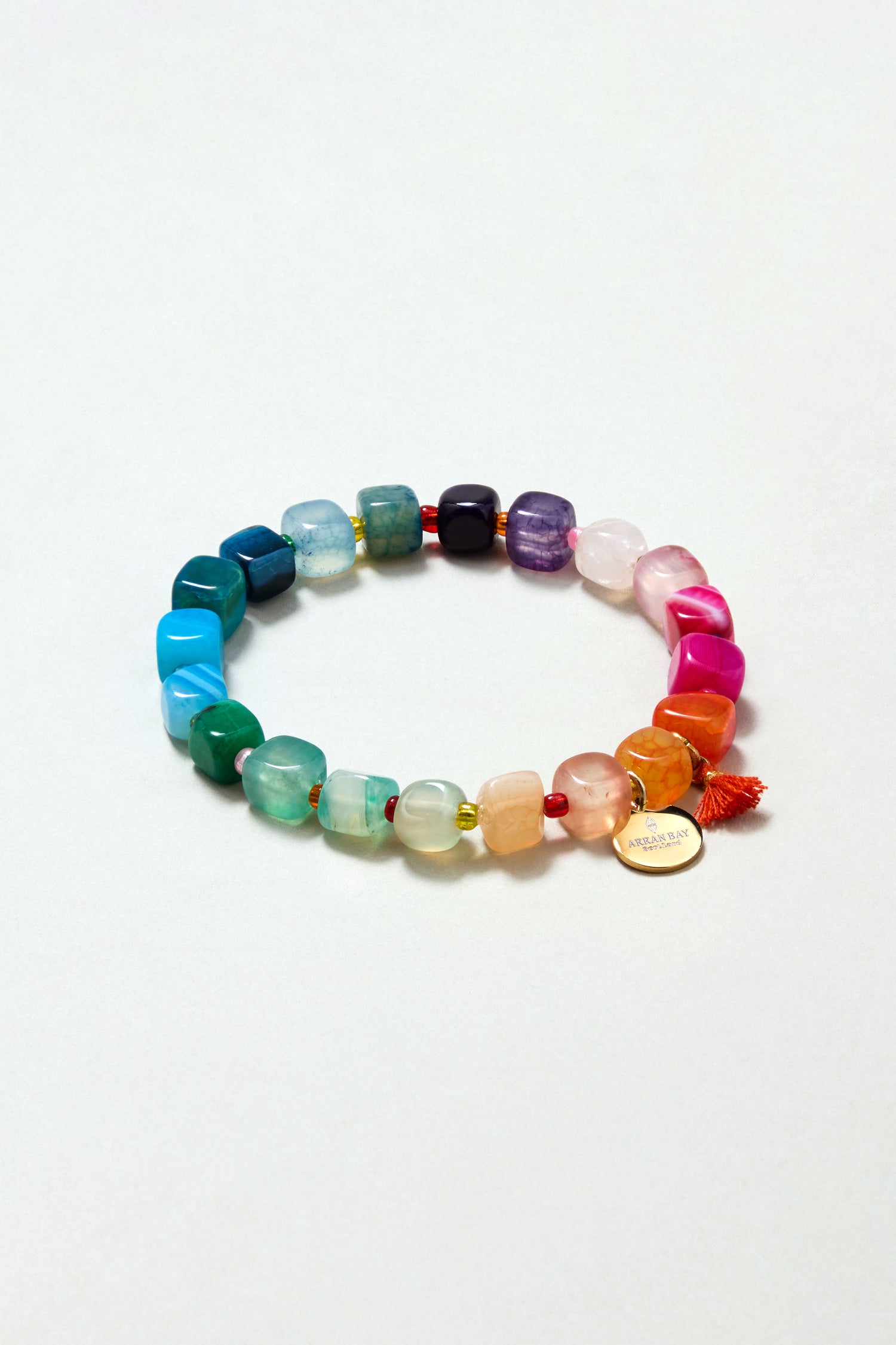 The Artisanal Rainbow Agate Bead Bracelet features multicolored irregular agate stones, a small orange tassel, and a round gold charm—handmade in Scotland and displayed against a light background.