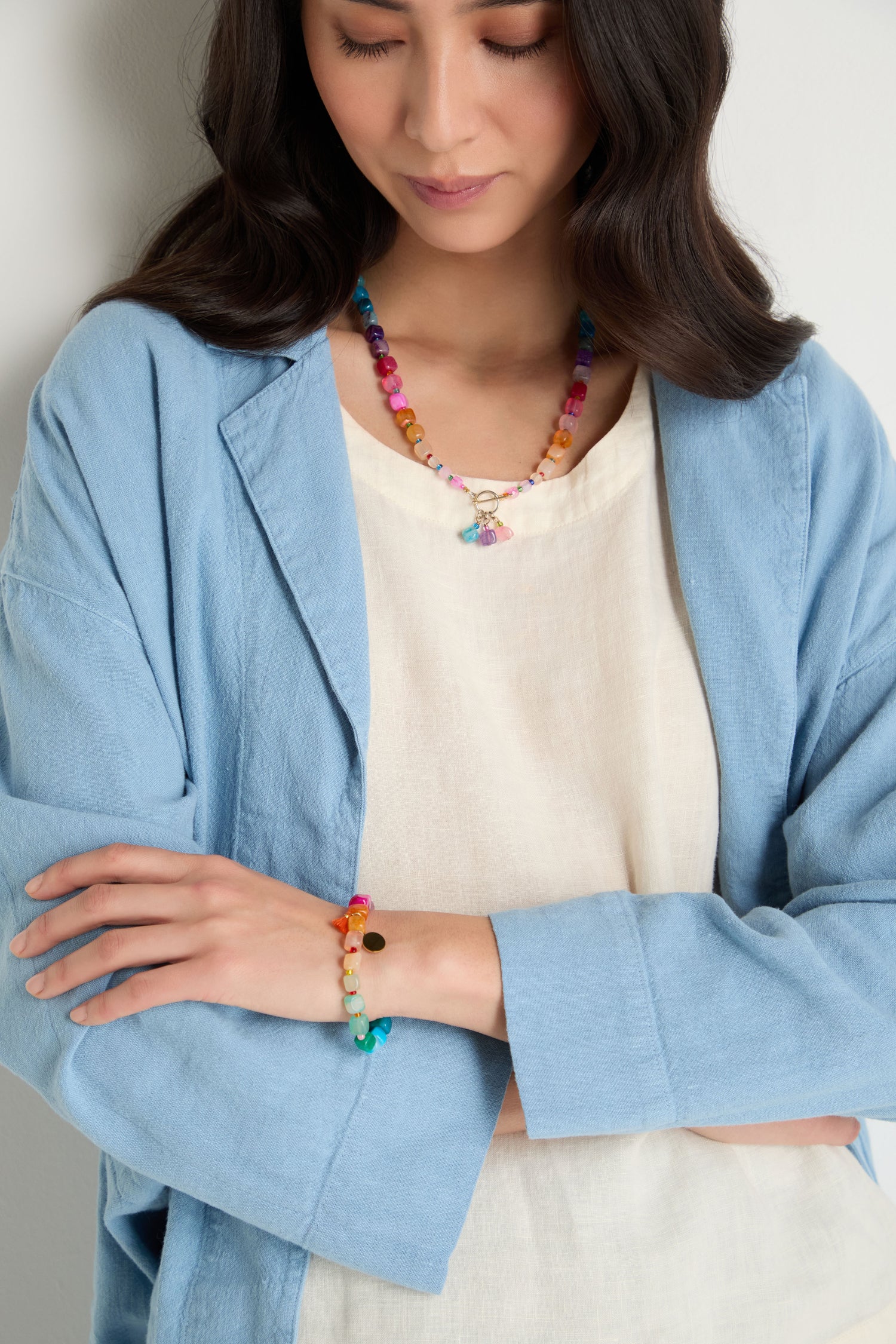 A woman in a light blue jacket and cream top displays the Artisanal Rainbow Agate Bead Bracelet, handmade in Scotland, along with a colorful beaded necklace, her arms crossed.
