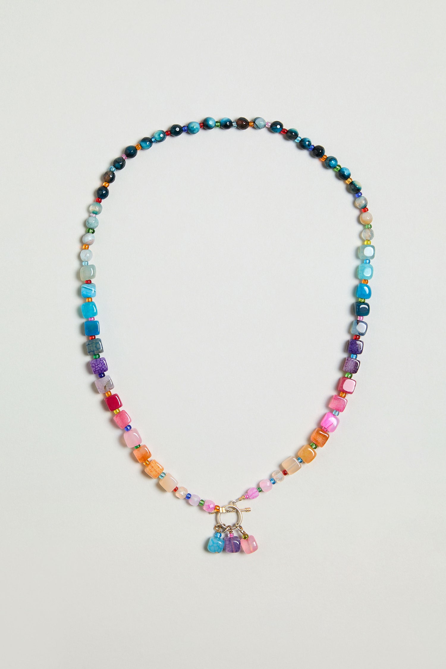 The Artisanal Rainbow Agate Toggle Necklace features multicolored agate beads in assorted shapes and sizes, accented with a cluster of vibrant charms at the center—handmade with care in Scotland.