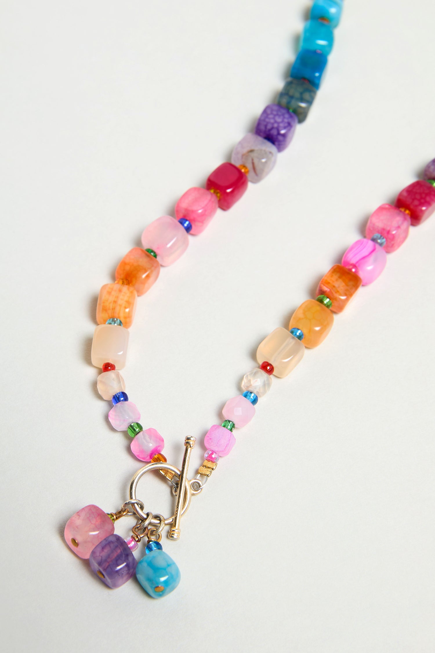 The Artisanal Rainbow Agate Toggle Necklace, handmade in Scotland with irregularly shaped rainbow agate beads and a gold toggle clasp, is displayed on a light background.