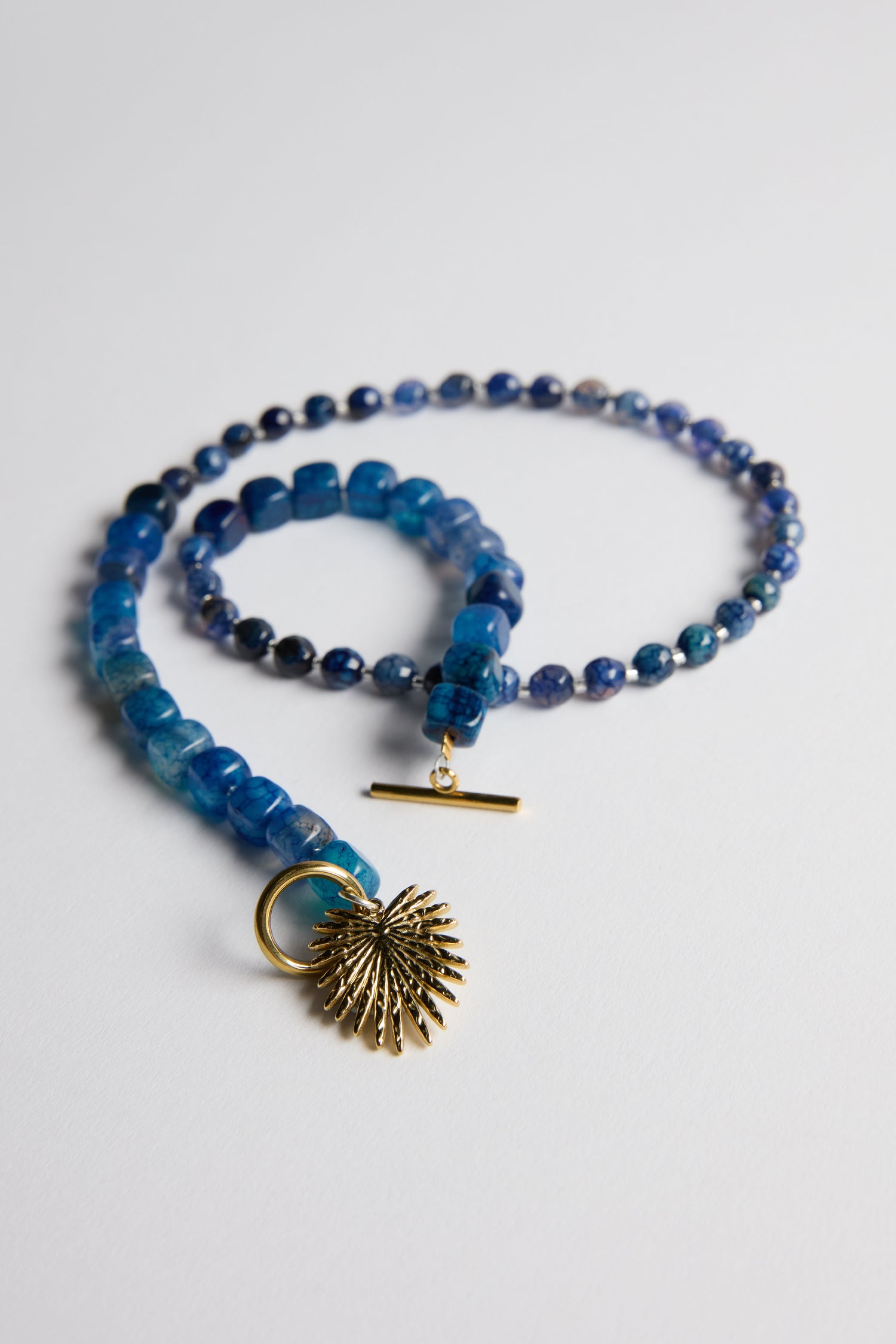 The Artisanal Blue Agate Palm Necklace, handmade in Scotland, showcases square blue agate stones, a gold toggle clasp, and a gold sunburst pendant—displayed elegantly on a white surface.