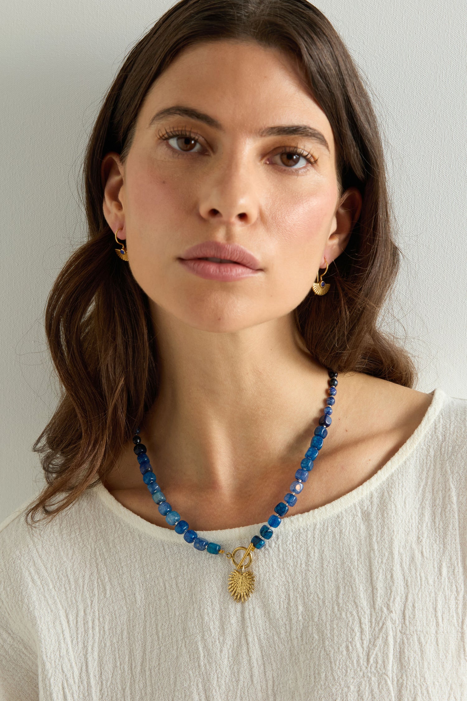 A woman with long brown hair wears a textured white top, small gold hoop earrings, and the Artisanal Blue Agate Palm Necklace—featuring a blue agate stone and gold pendant—while standing against a light background.