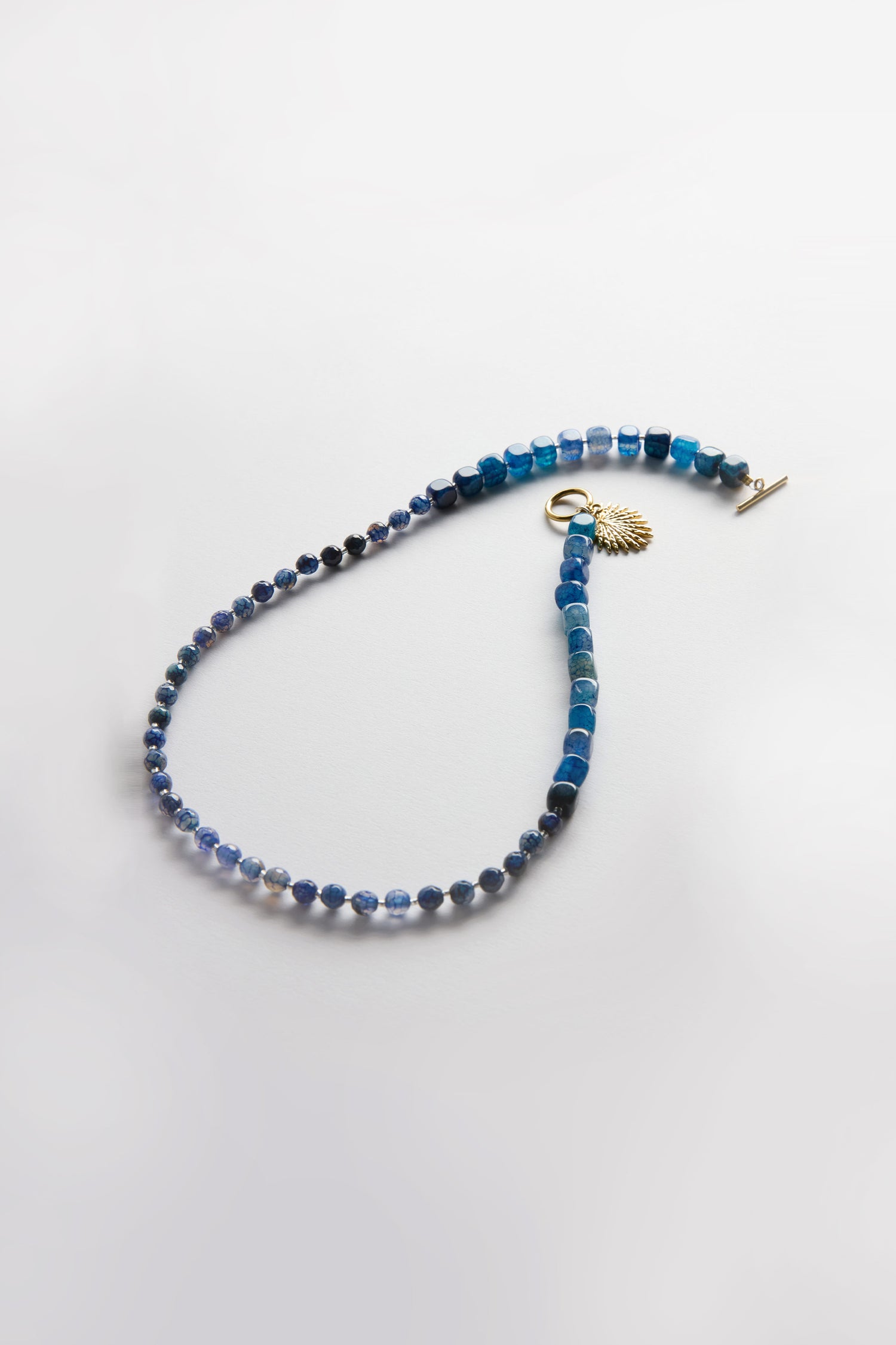 The Artisanal Blue Agate Palm Necklace from Arran Bay Jewellery features blue agate beads, a gold clasp, and a gold-plated palm charm, shown on a light background.