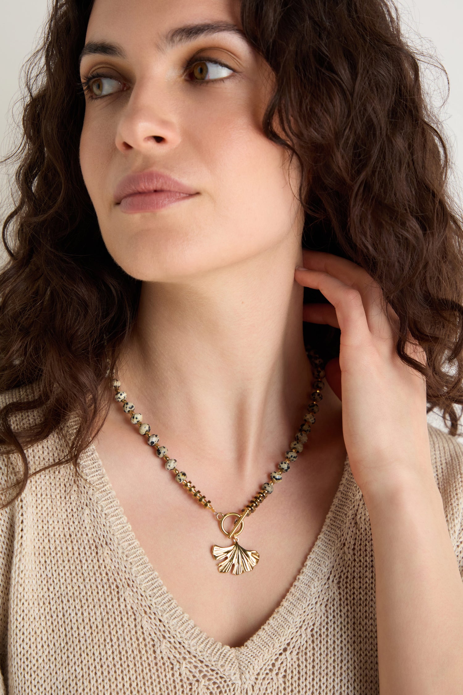 A woman with curly brown hair wears a beige knit top and the Dalmatian Stone Ginkgo Leaf Necklace, looking slightly to the side with one hand near her neck.