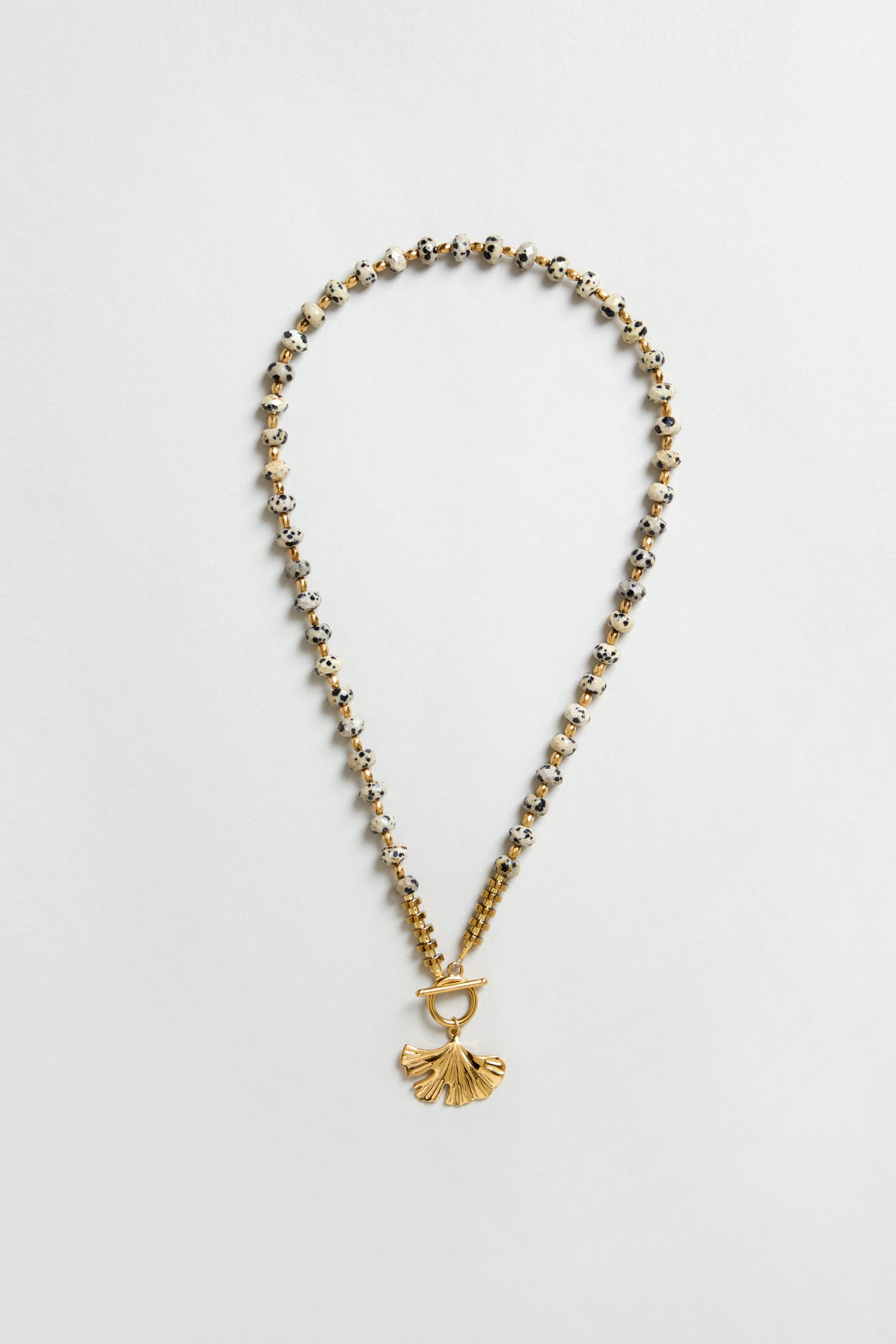 The Dalmatian Stone Ginkgo Leaf Necklace features gold and silver beads, a textured ginkgo leaf pendant, and a toggle clasp, displayed on a flat neutral background.