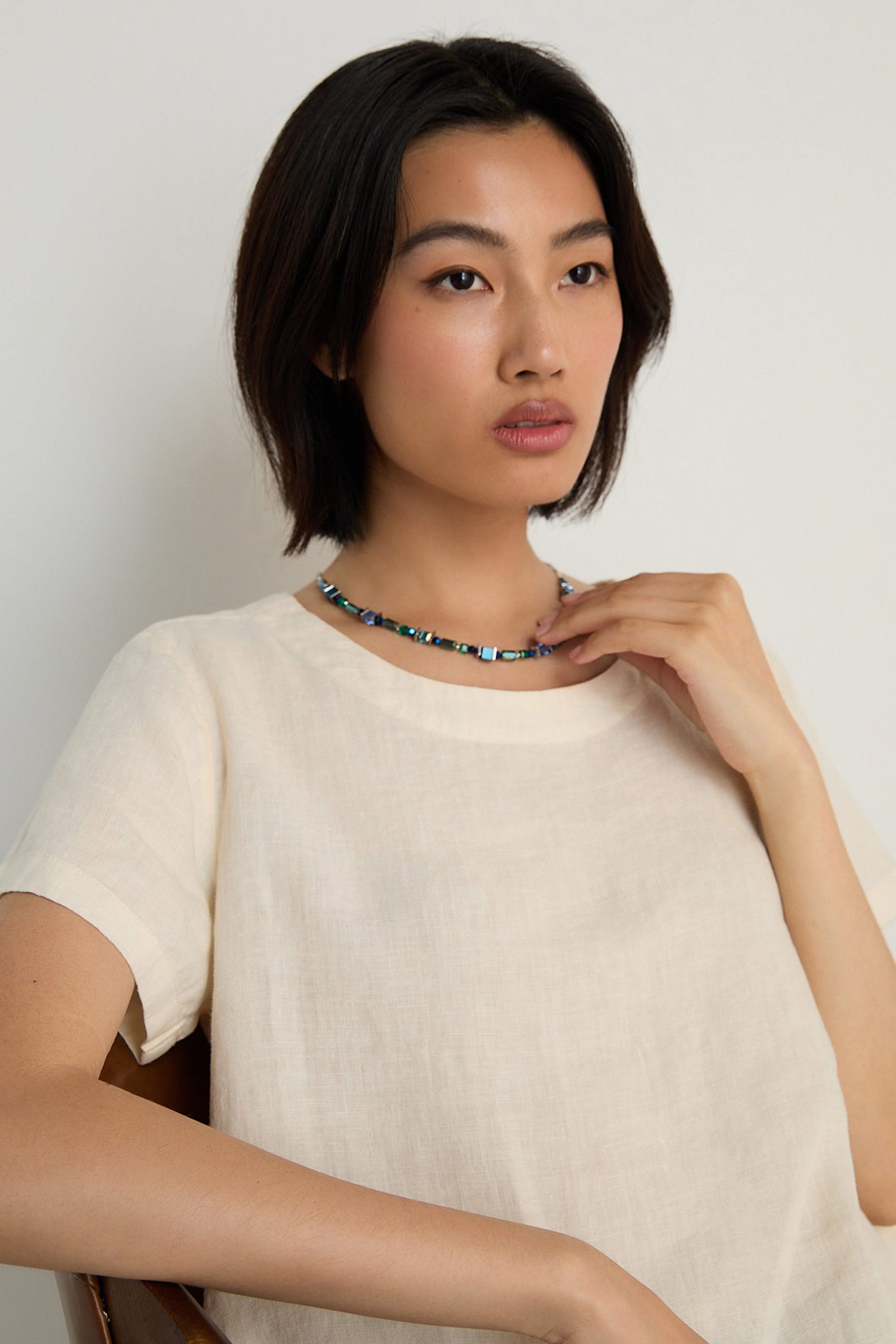 A person with short dark hair, wearing a light-colored top and the Blue Hematite Necklace, sits against a plain background looking slightly to the side.