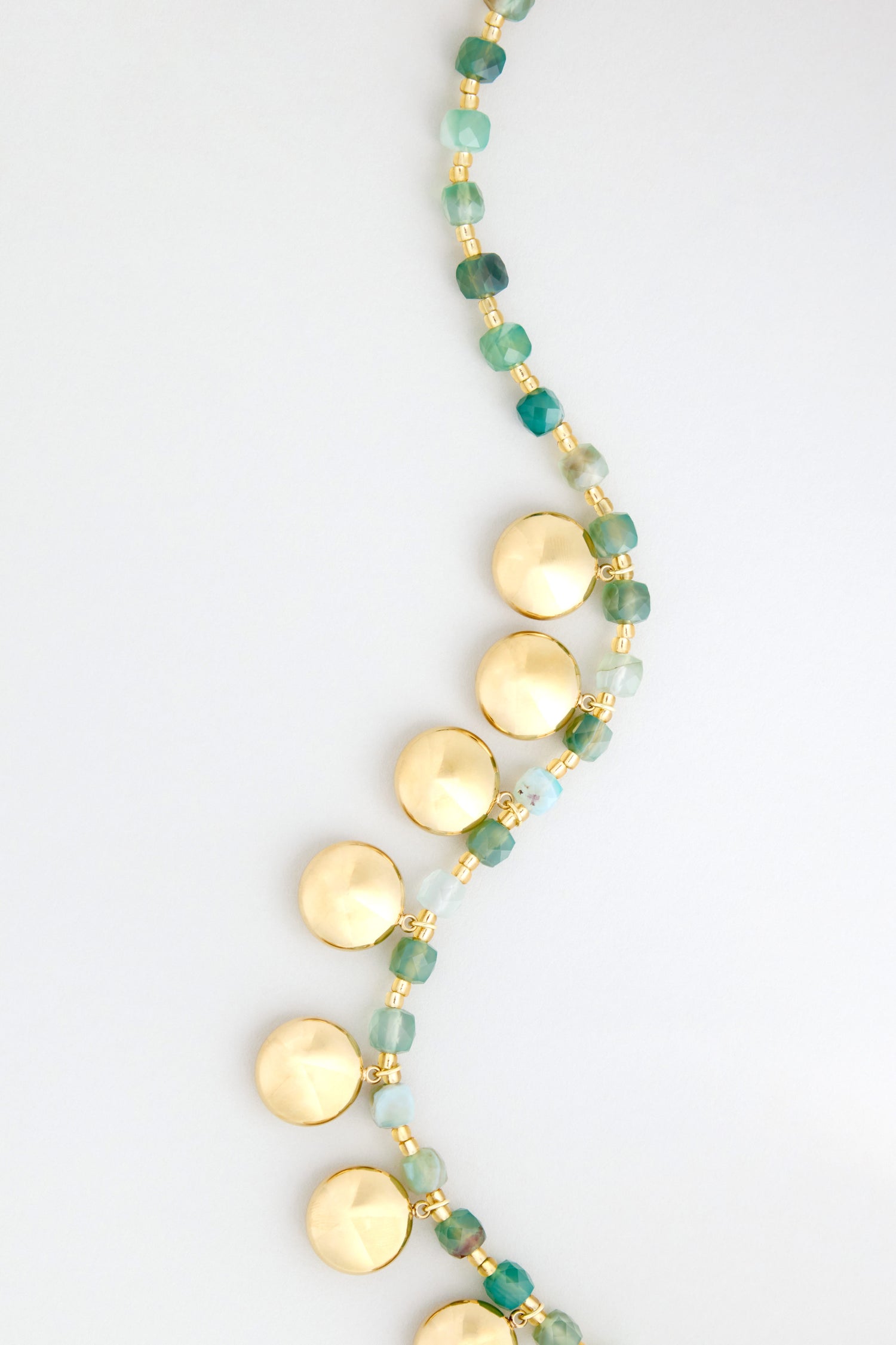 The Faceted Green Agate Charm Necklace showcases green-blue beads and round gold discs arranged in a curved line, set against a light background.