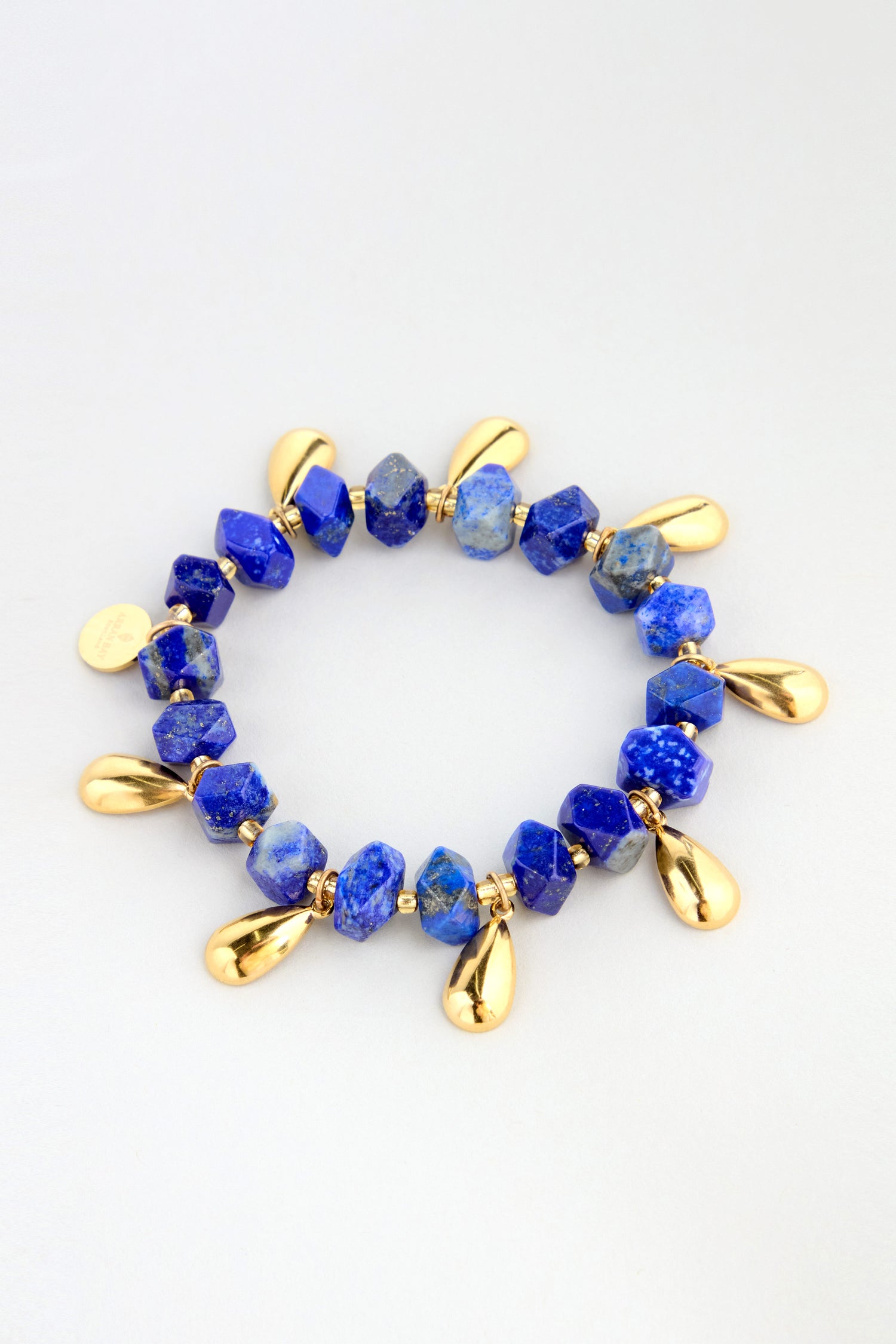 The Lapis Lazuli Teardrop Bracelet features blue stone beads and gold teardrop-shaped charms, beautifully arranged in a circle on a light background.