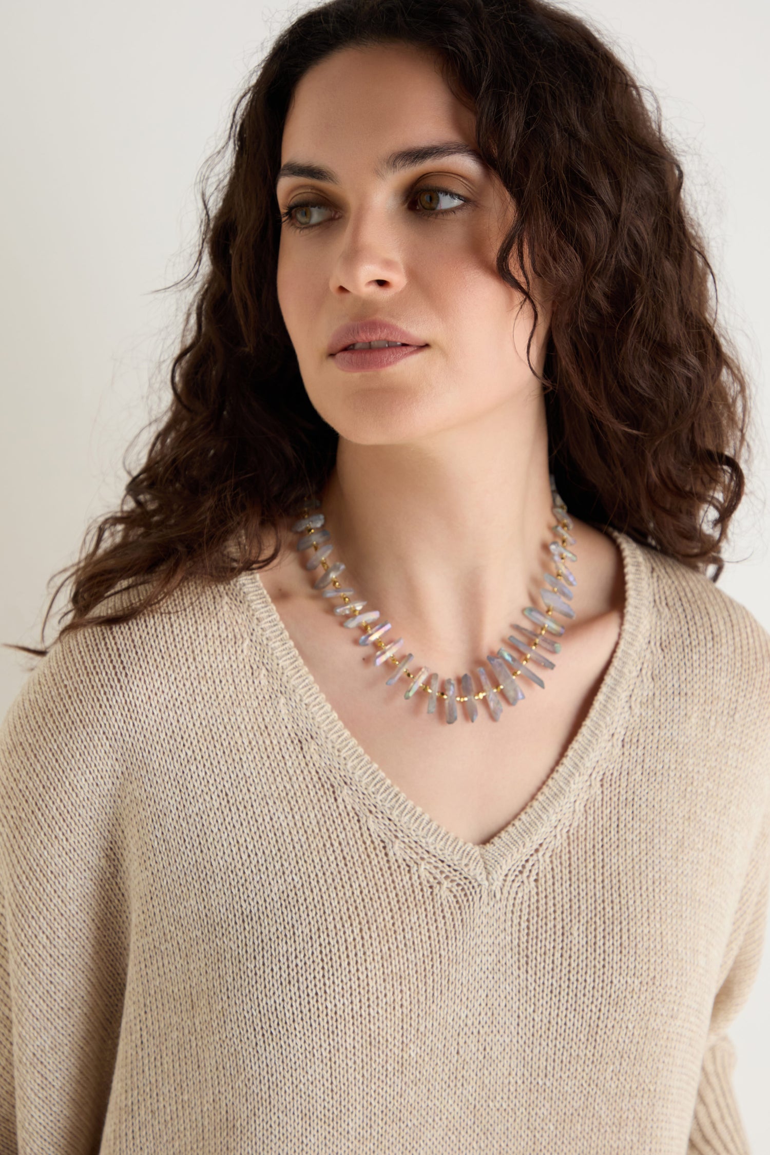 A woman with wavy brown hair looks to the side against a neutral background, wearing a beige knit sweater and the Slate Quartz Necklace.