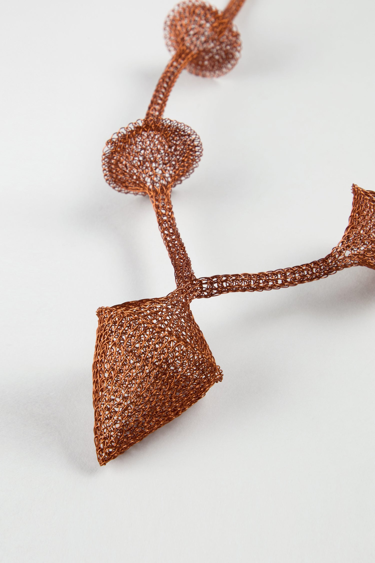 The Hand Crocheted Snowdrop Necklace features intricately woven, copper-colored wire in geometric circular and diamond shapes, beautifully displayed on a white background.