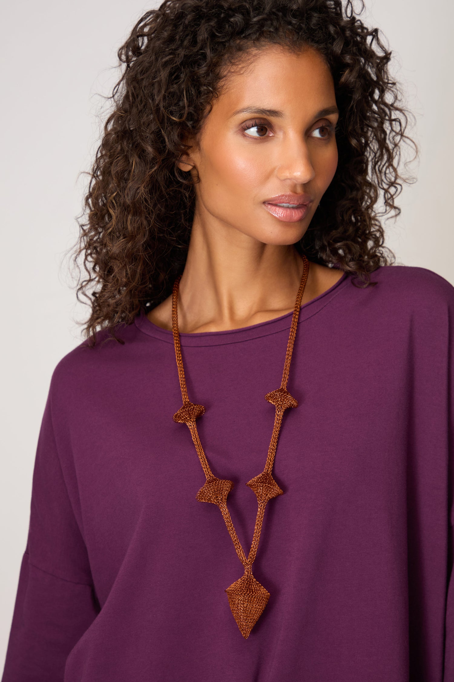A woman with curly hair wears a purple top and the Hand Crocheted Snowdrop Necklace, gazing slightly to the side against a plain background.