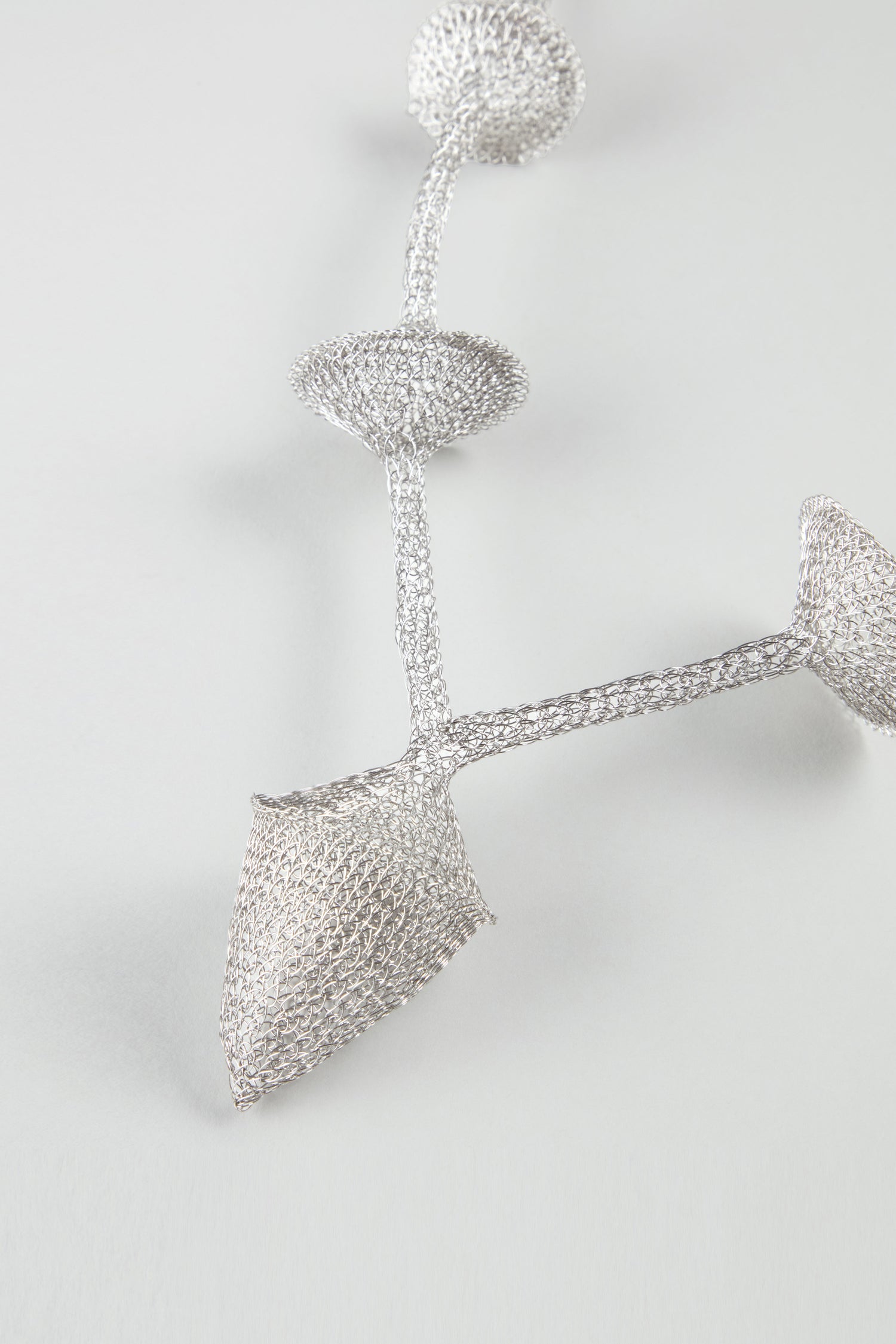 A close-up of the Hand Crocheted Snowdrop Necklace reveals abstract, interconnected vessel-like forms, evoking contemporary jewelry design against a plain light background.