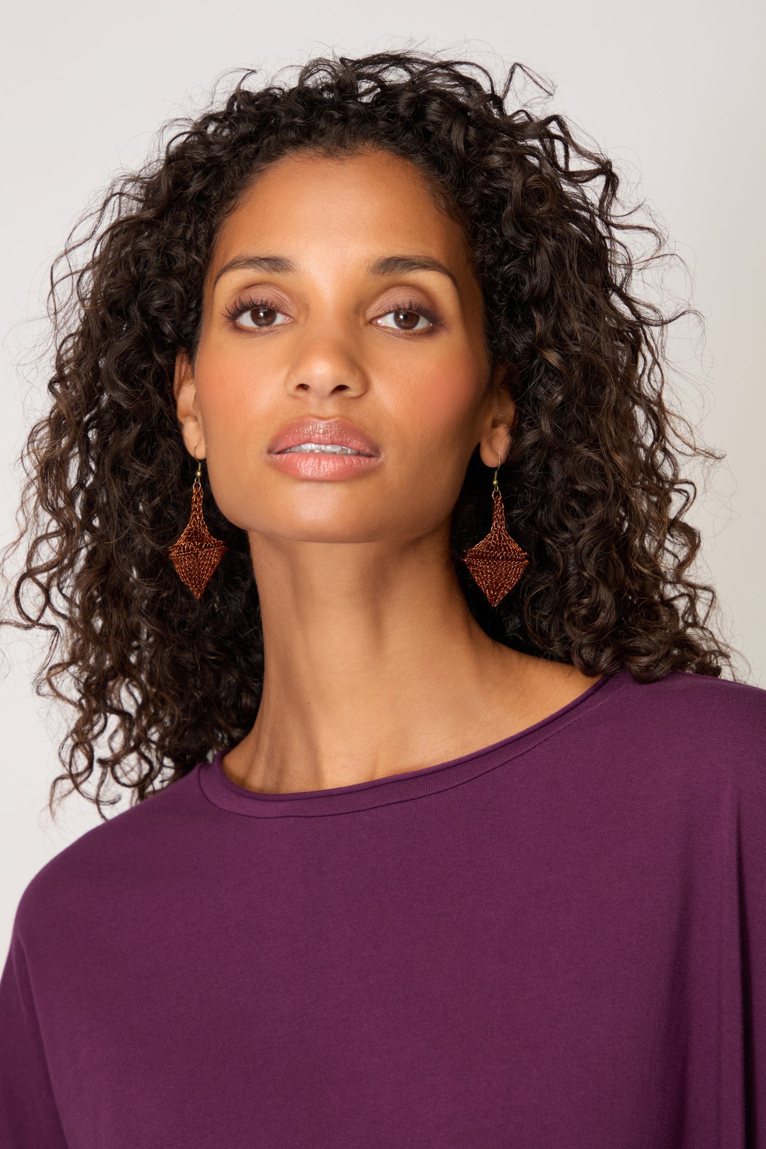 A woman with curly hair wears a purple top and showcases Hand Crocheted Earrings, gazing directly at the camera against a plain background.