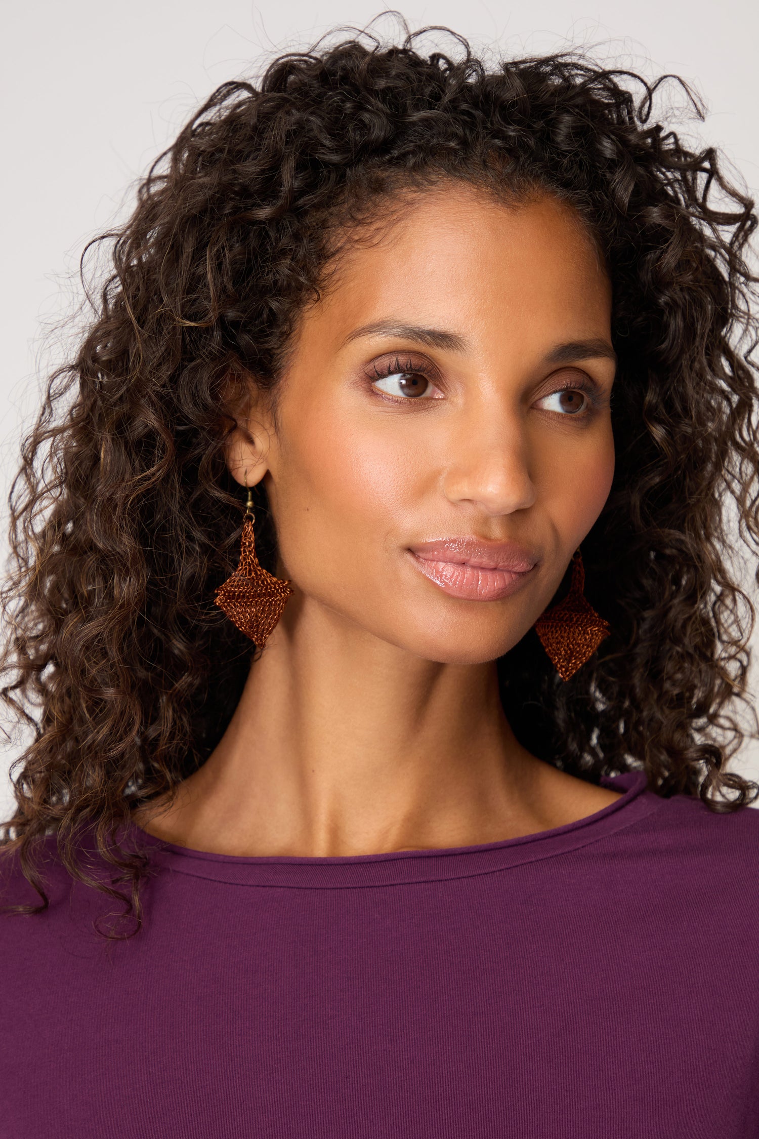 A woman with curly hair, wearing a purple top and Hand Crocheted Earrings, glances slightly to the side against a plain background.