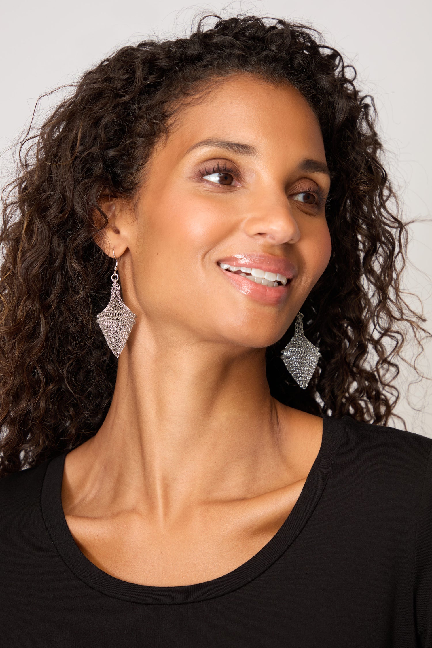 Woman with curly hair smiles and looks to the side against a plain background, wearing a black top and Hand Crocheted Earrings.