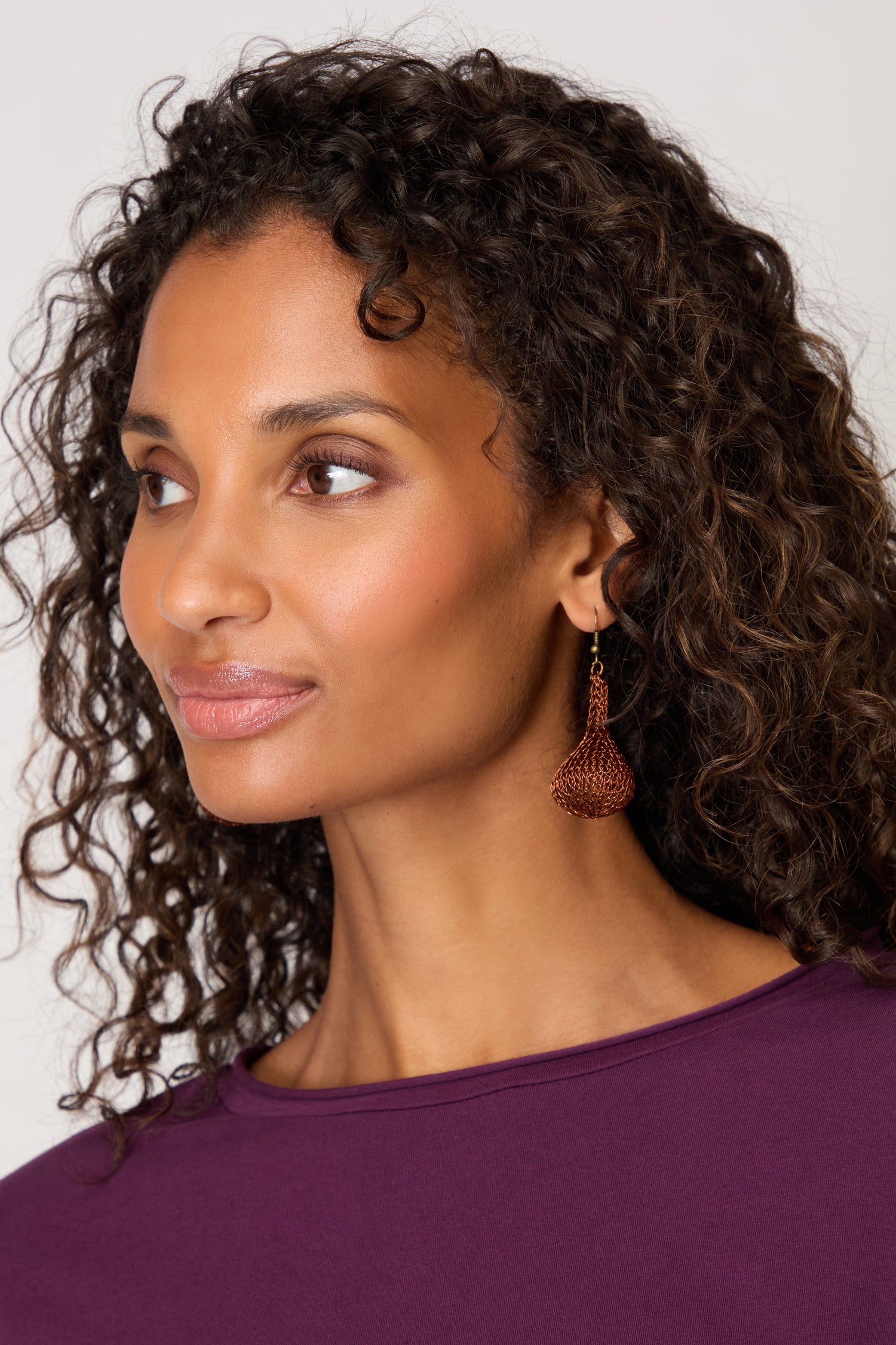 A woman with curly hair in a purple top wears a single brown Hand Crocheted Teardrop Earring, highlighting its artisanal design as she gazes to the side against a simple background.