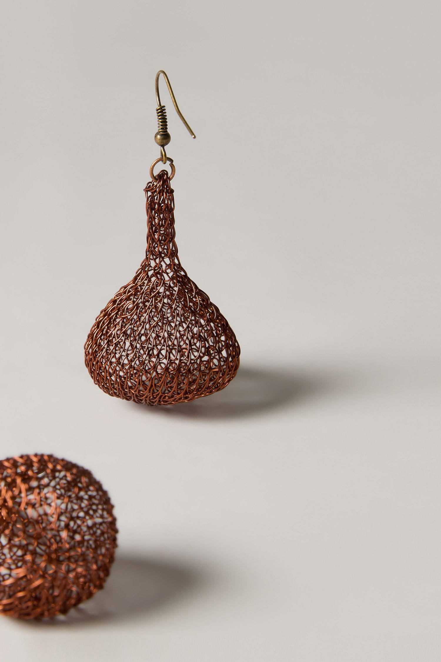 Hand Crocheted Teardrop Earrings, featuring a handmade brown wire mesh with a bulbous shape, hang against a plain light background. Another earring appears blurred in the foreground, highlighting their artisanal and lightweight design.