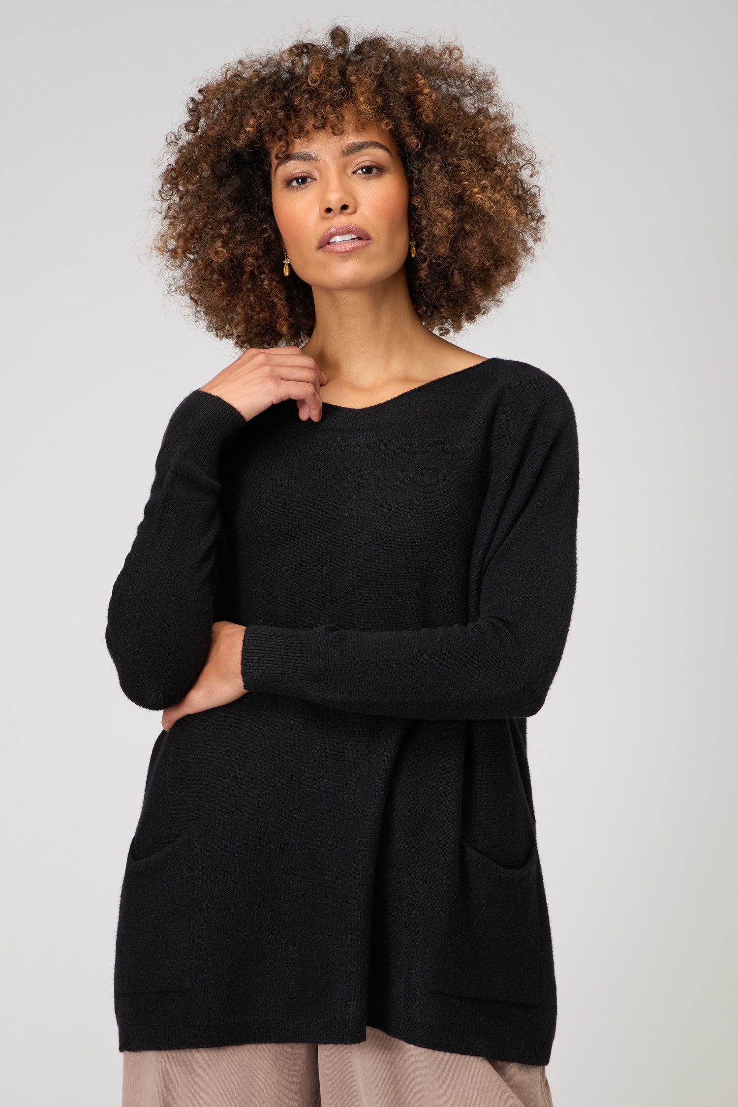 A woman with curly hair models the Two Pocket Oversize Knit, a black supersoft long-sleeve sweater, paired with light-colored pants against a plain light background.