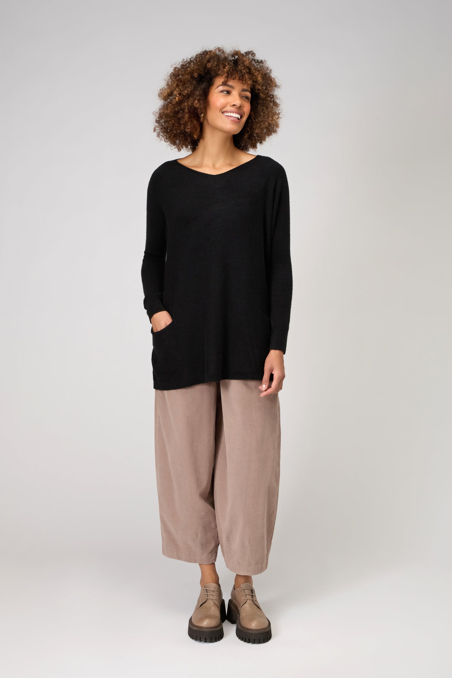 A person stands against a plain background, smiling with hands in pockets, wearing the Two Pocket Oversize Knit in black, paired with beige wide-leg pants and tan shoes.