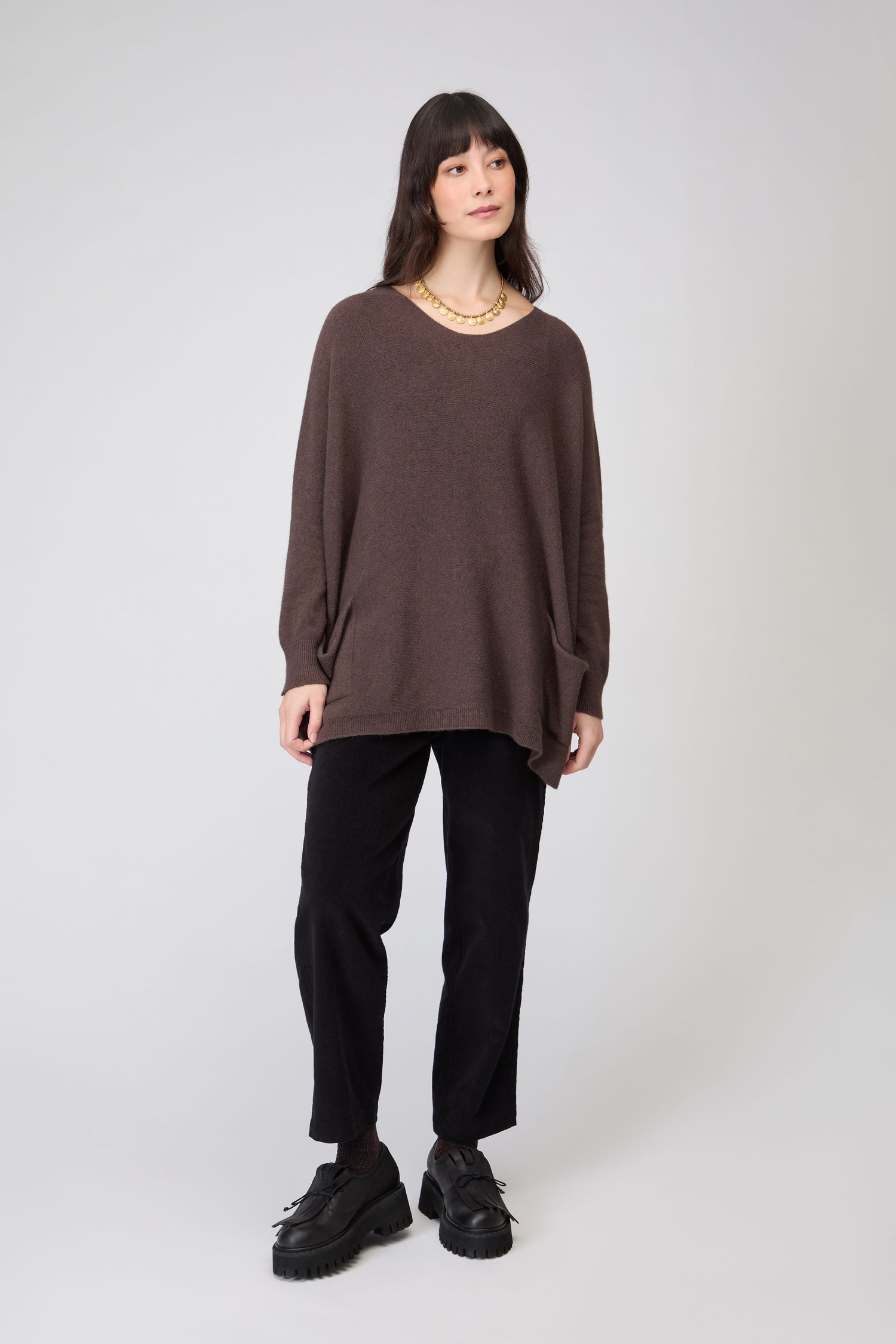 A woman stands against a plain background wearing the Two Pocket Oversize Knit jumper in brown, paired with black pants, black shoes, and a gold necklace.