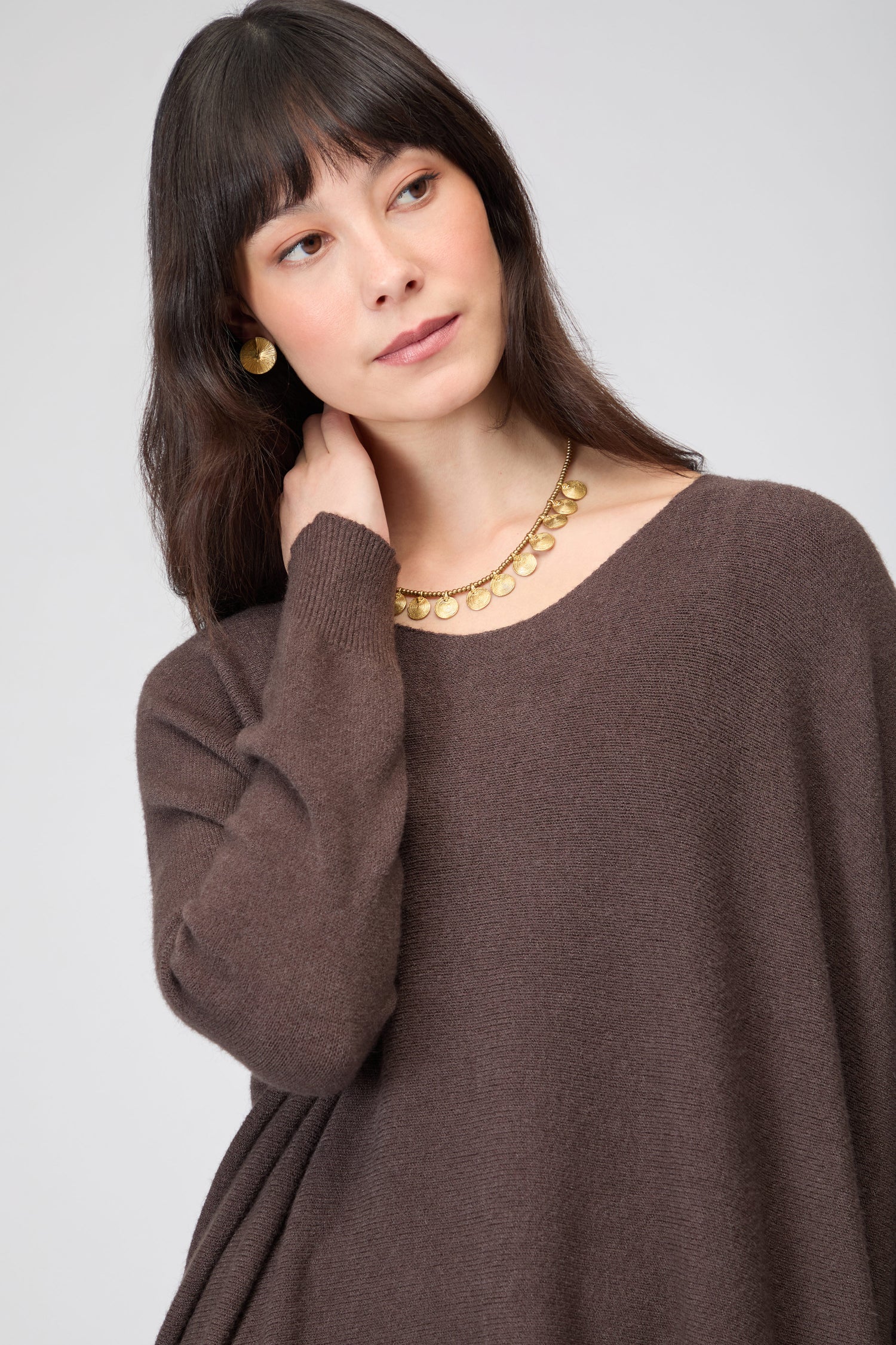 Woman with long dark hair wears the Two Pocket Oversize Knit in brown, styled with gold earrings and a gold necklace, looking slightly to the side against a plain background.