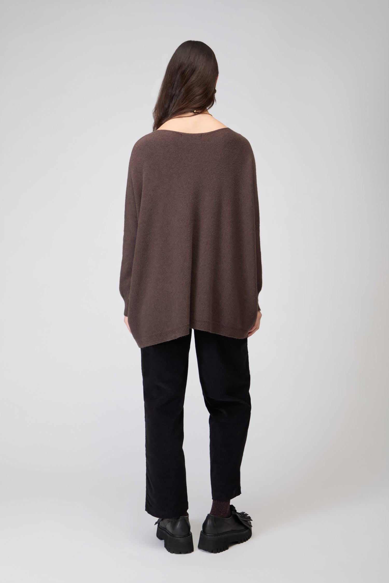 A person faces away from the camera, wearing a brown Two Pocket Oversize Knit jumper, black pants, and black shoes against a plain light background.