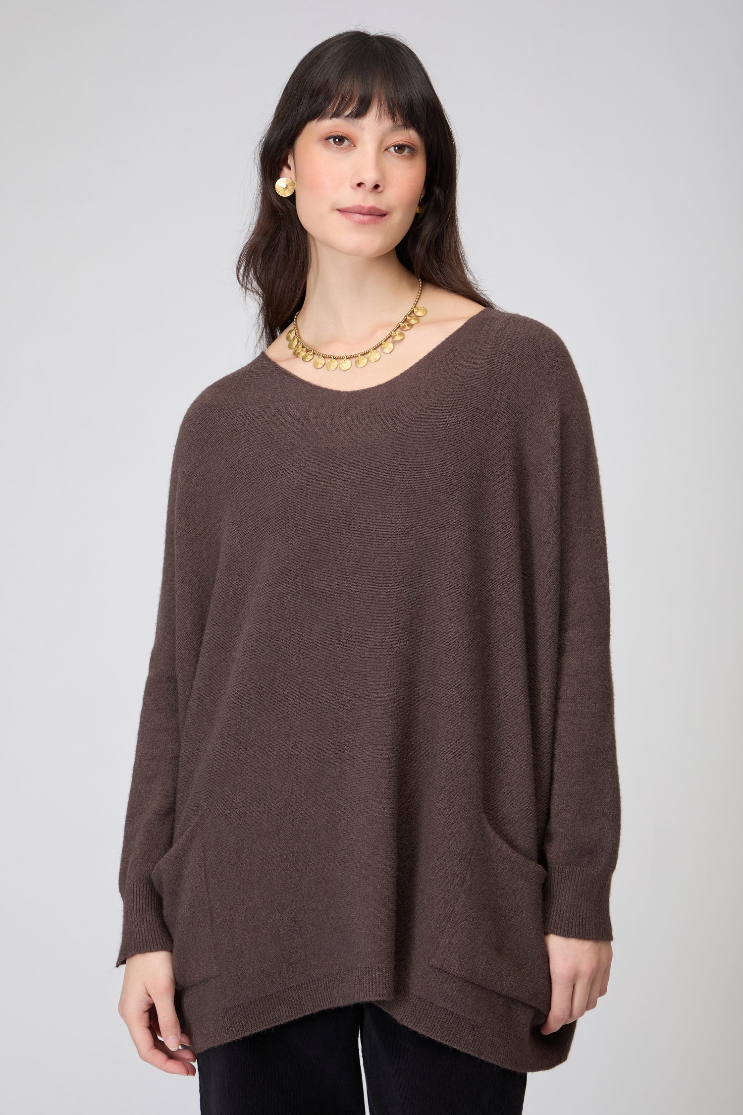 A woman wears the Two Pocket Oversize Knit in brown, paired with black pants, gold earrings, and a gold necklace, standing against a plain light gray background.