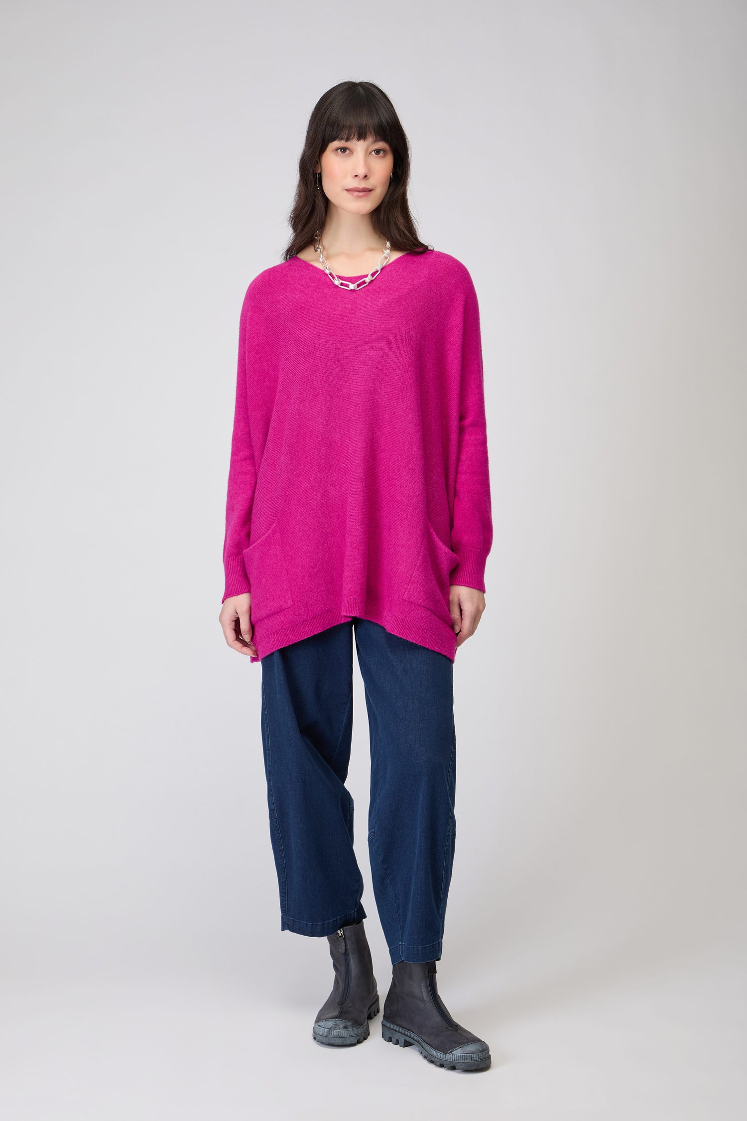 A woman faces forward, wearing the Two Pocket Oversize Knit in bright pink, paired with dark blue pants and gray boots against a plain light background.