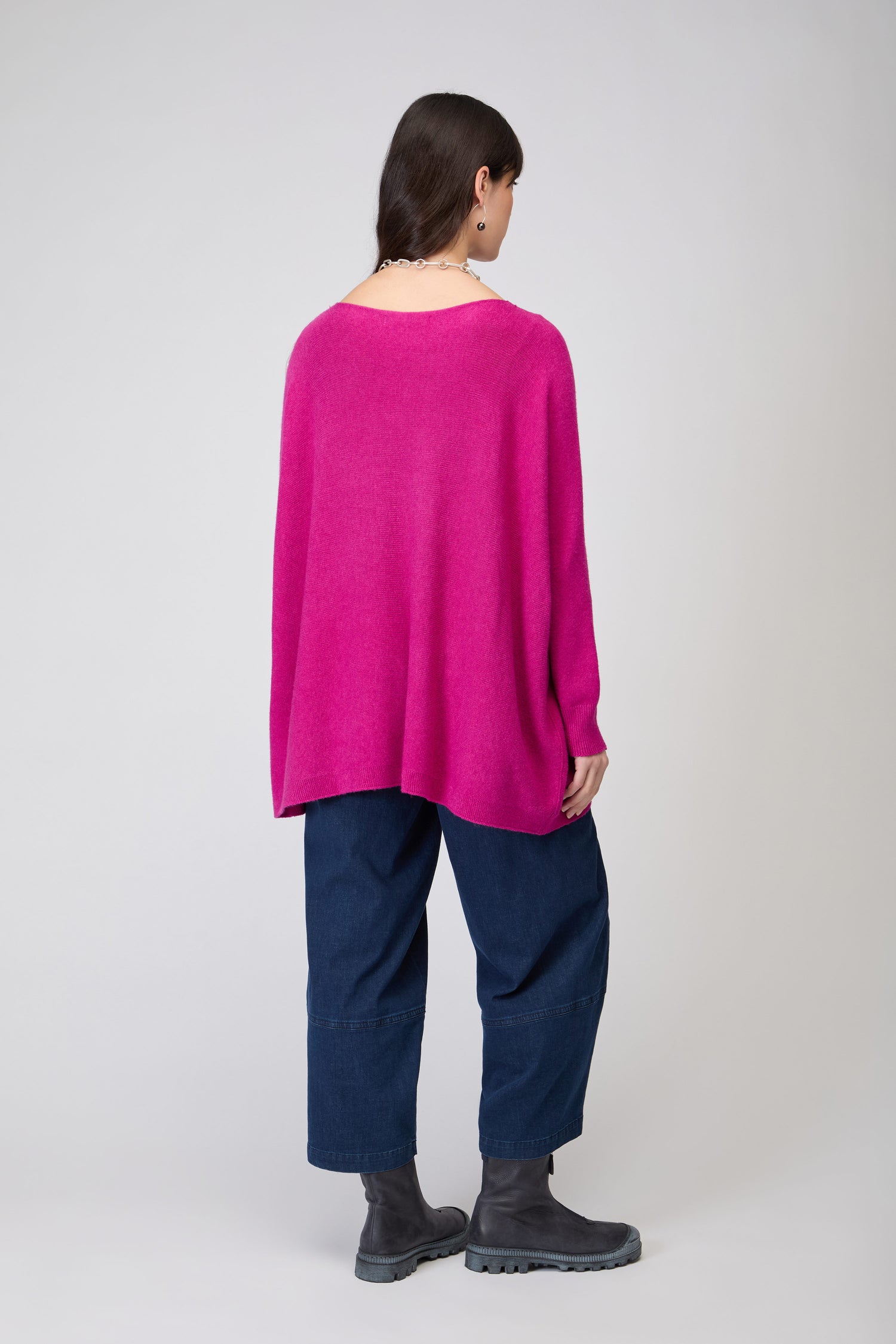 A person stands facing away, wearing the Two Pocket Oversize Knit in bright pink, paired with dark blue wide-leg pants and gray boots against a light neutral background.