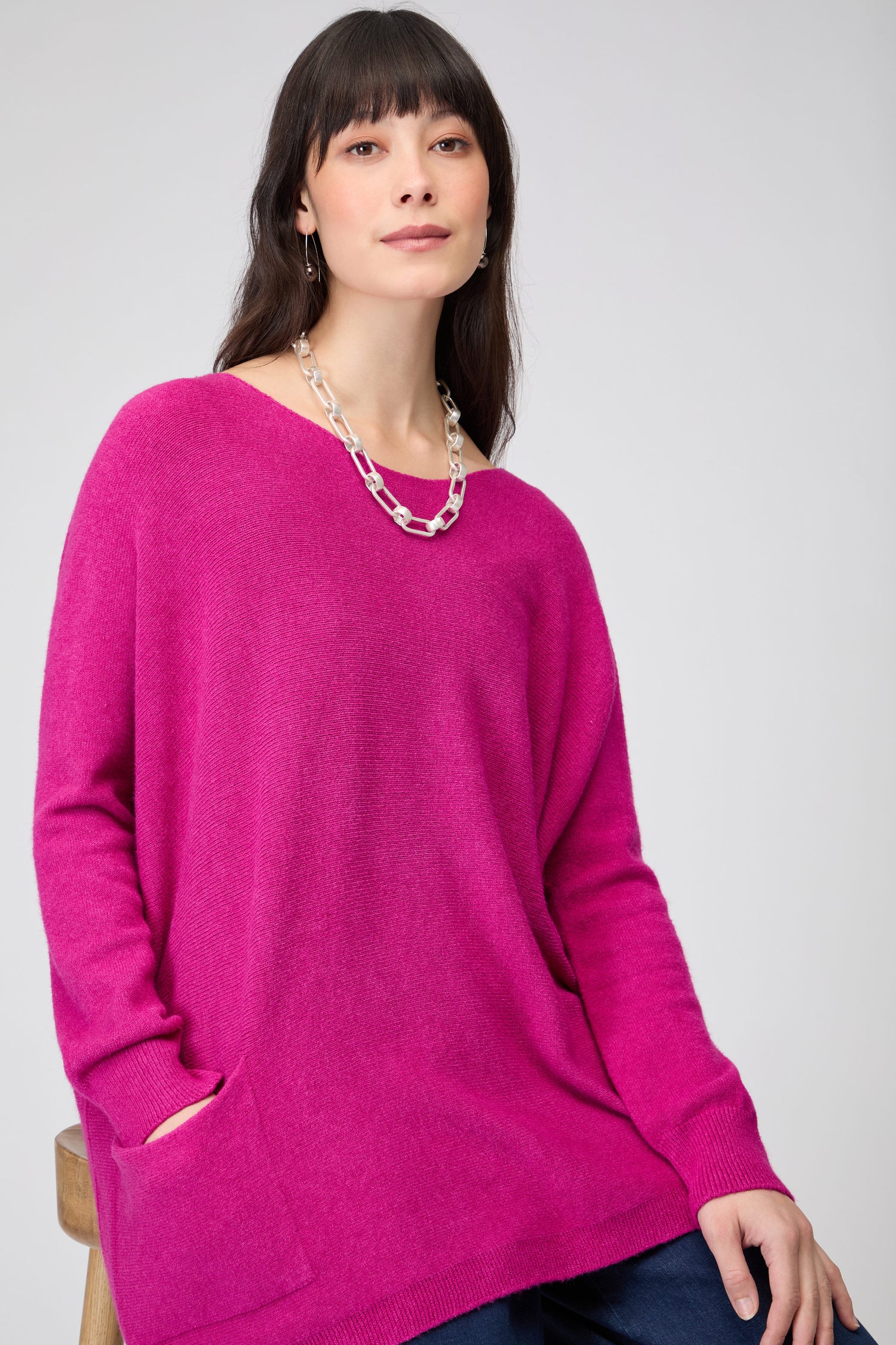A woman sits on a stool against a plain background, wearing the Two Pocket Oversize Knit in bright pink with long sleeves, paired with a silver chain necklace and dark pants.