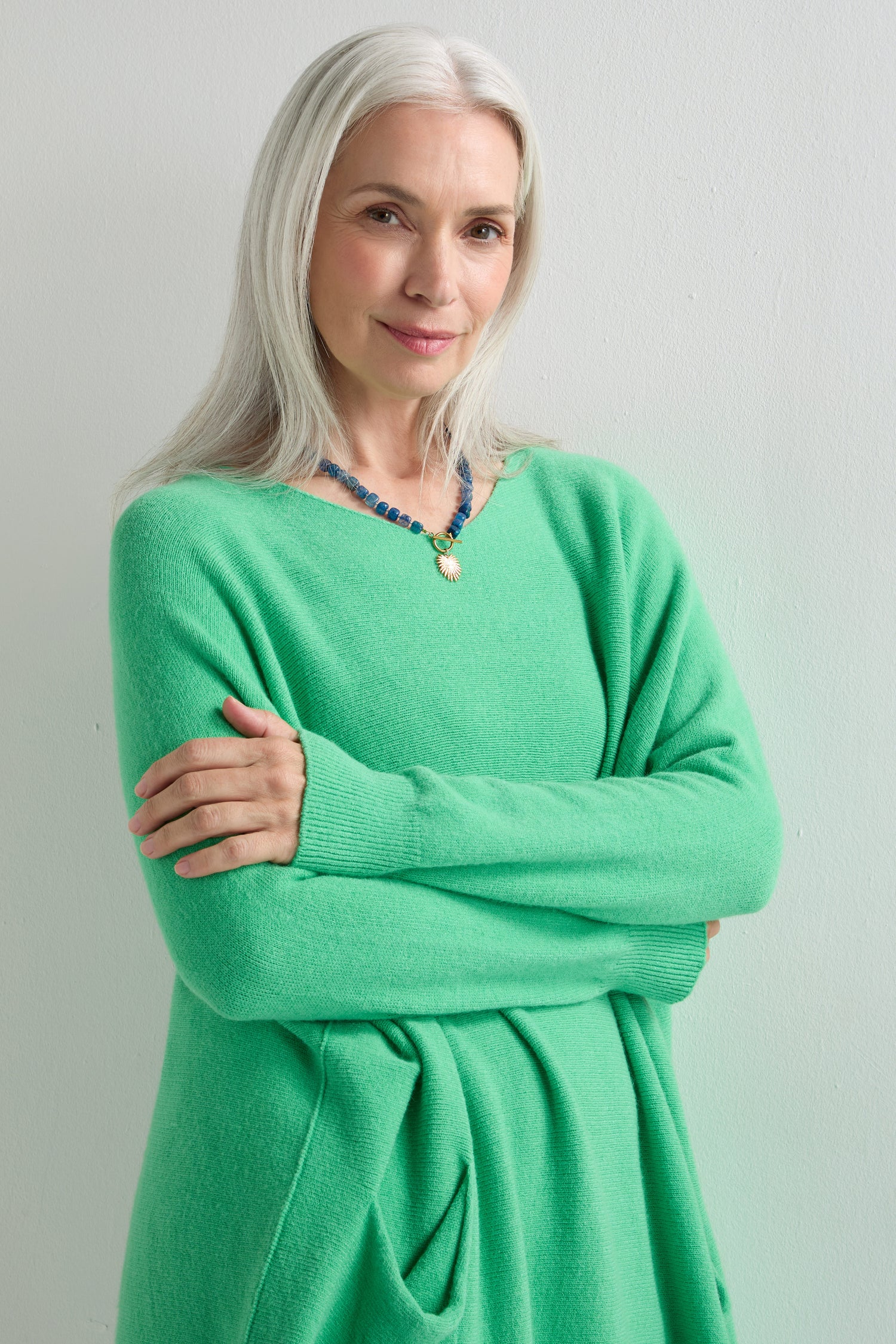 A stylish woman with long gray hair stands against a white wall, wearing the Two Pocket Oversize Knit in bright green and a blue beaded necklace, arms crossed and smiling slightly.