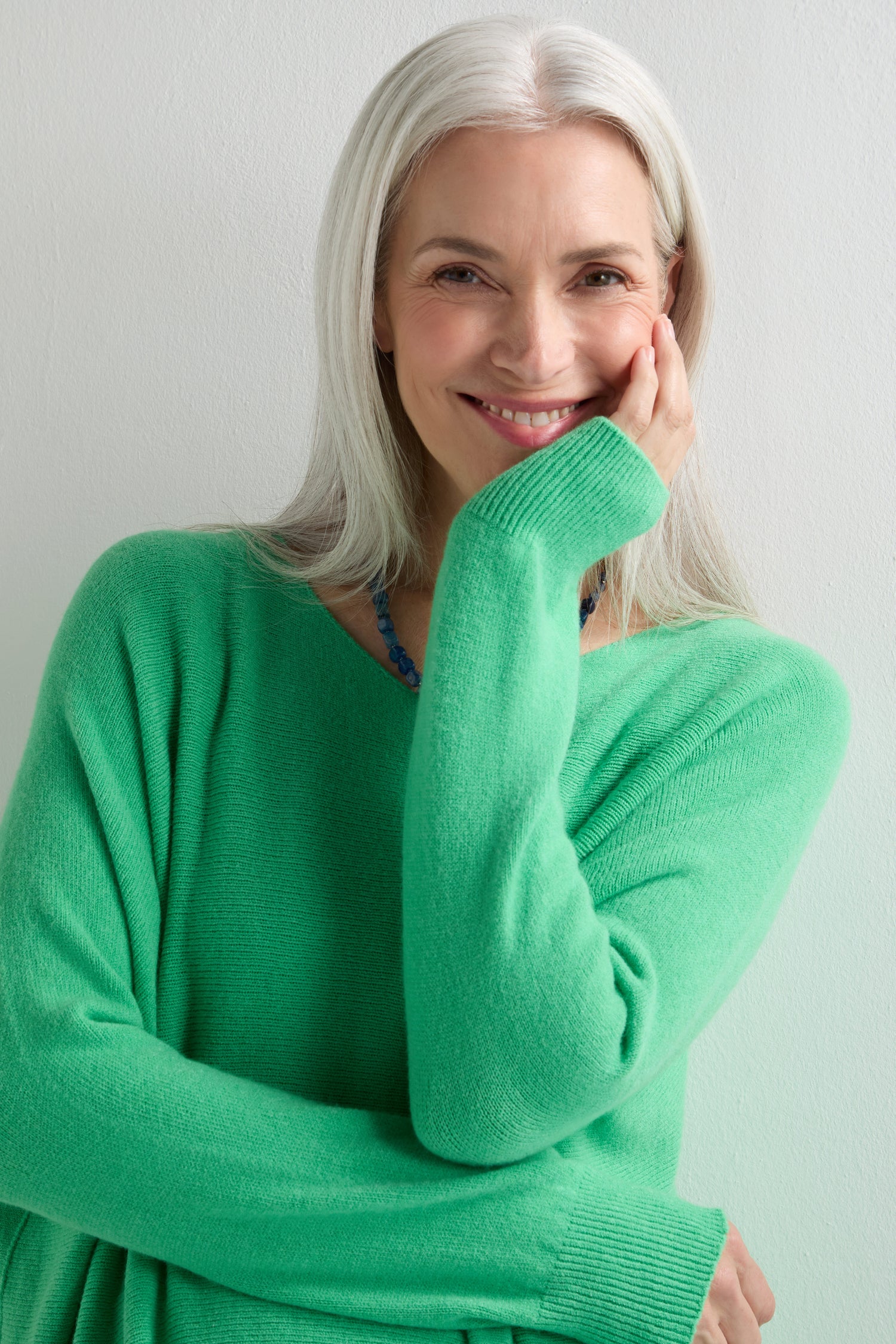 A woman with long gray hair in an oversized green Two Pocket Oversize Knit sweater smiles, resting her hand on her cheek against a light background—an effortless style from Amazing Woman.