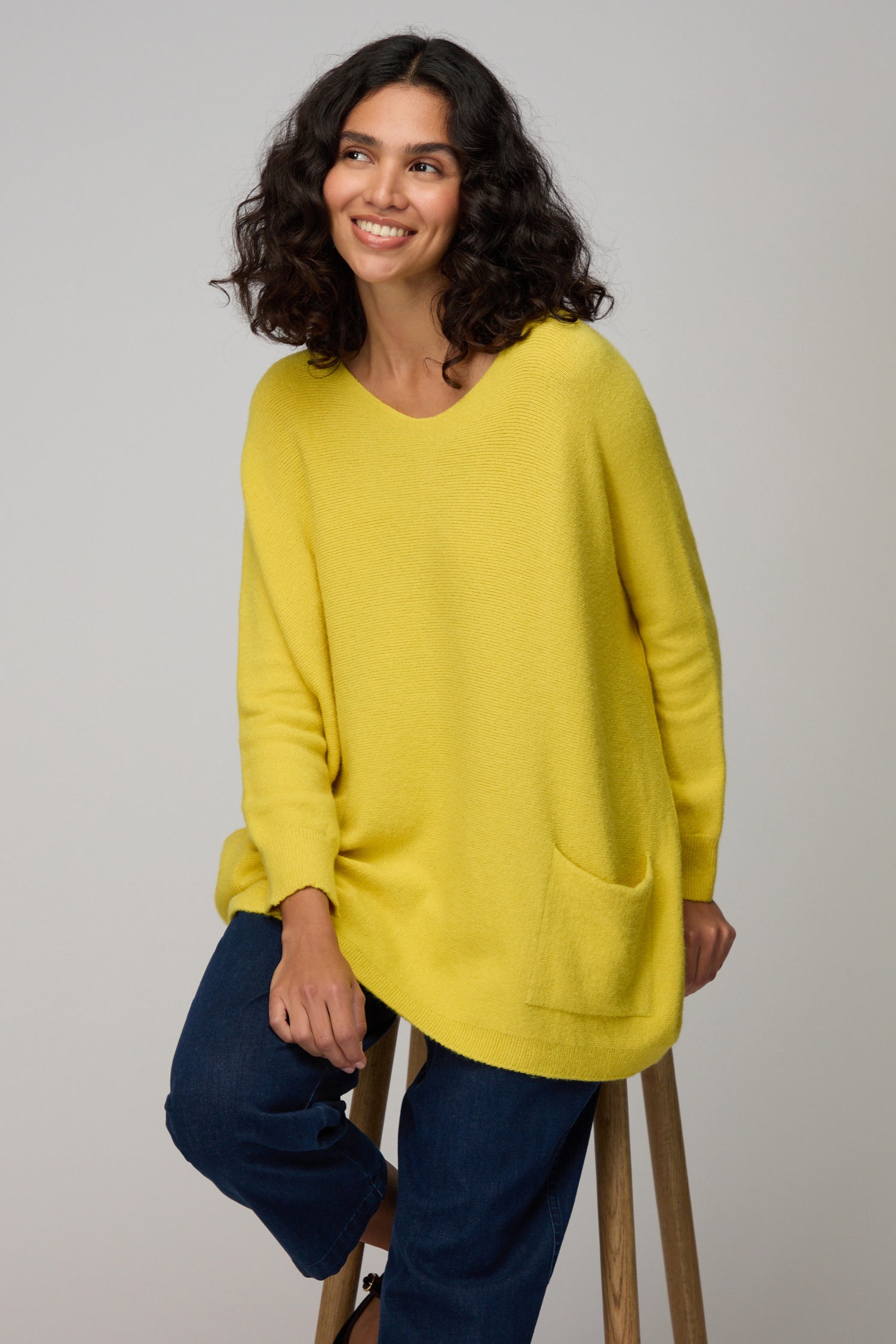 Woman with curly hair wearing the Two Pocket Oversize Knit in yellow and dark jeans sits on a wooden stool, smiling against a light gray background—an effortlessly chic style from Amazing Woman’s supersoft knits collection.
