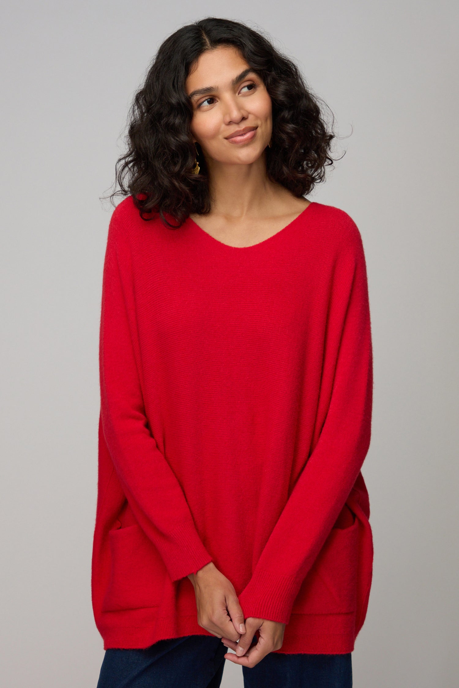A woman with curly hair models the Two Pocket Oversize Knit in red—an effortless look from Amazing Woman—paired with dark pants against a plain gray background.