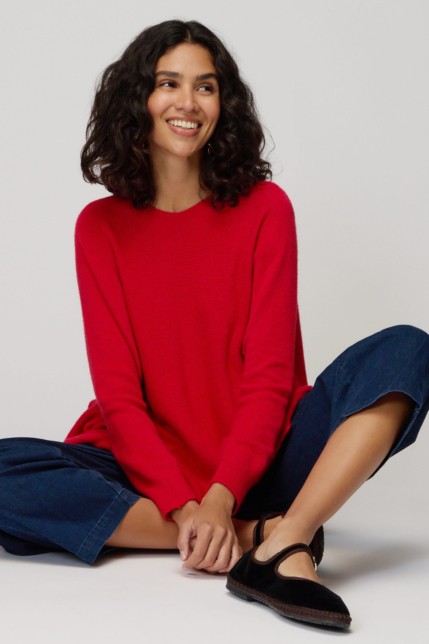 A woman with curly dark hair sits cross-legged on the floor, smiling and looking aside, wearing a red Two Pocket Oversize Knit sweater, blue pants, and black shoes against a plain background.
