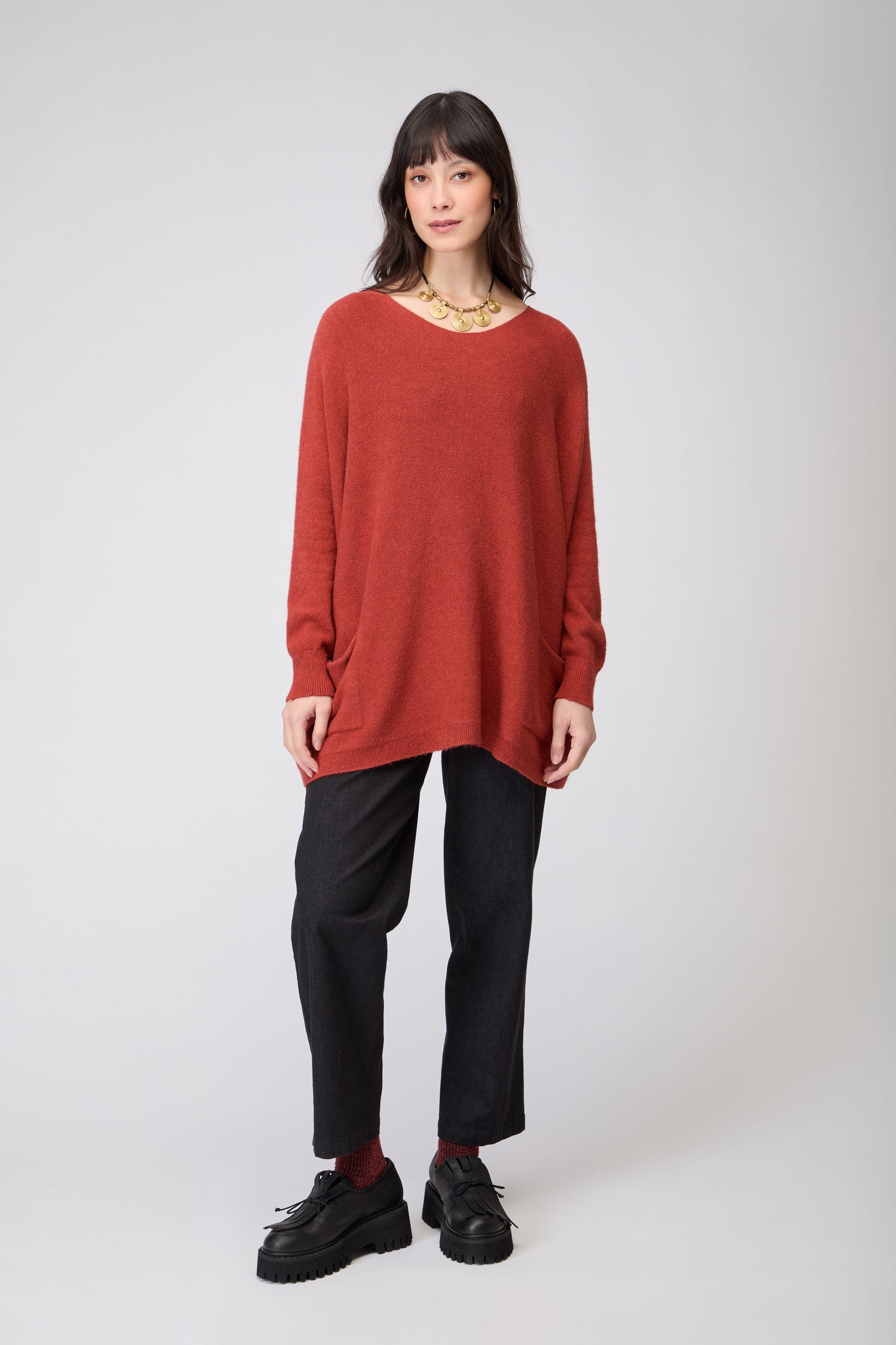 A person stands facing forward against a plain light background, wearing the Two Pocket Oversize Knit in red, dark pants, black shoes, and a gold necklace.