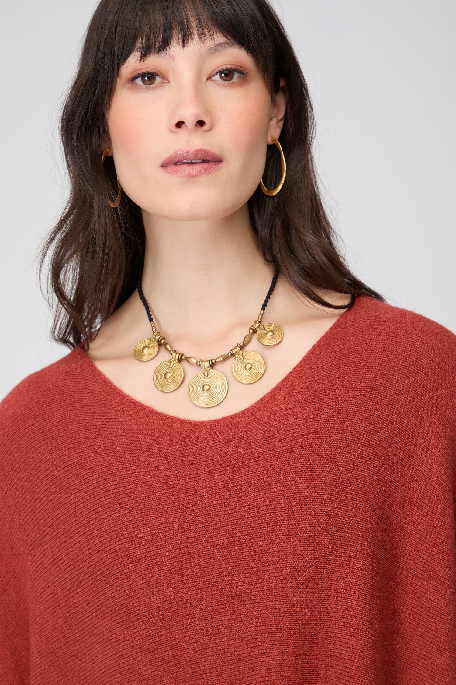 A woman models the Two Pocket Oversize Knit in rust, styled with gold hoop earrings and a chunky gold pendant necklace, looking directly at the camera against a plain background.