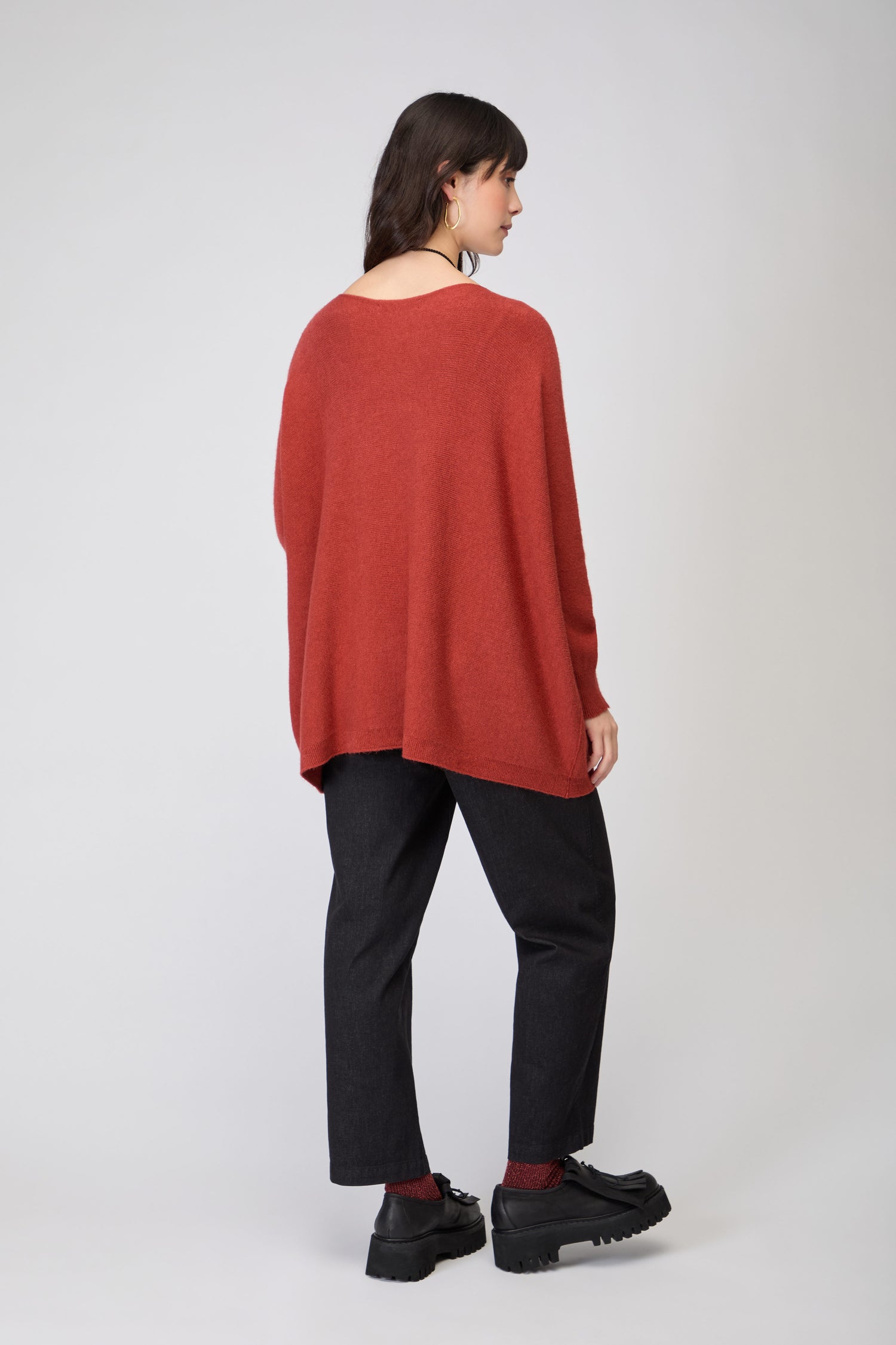 A woman stands against a plain background, facing away. She wears the Two Pocket Oversize Knit in red, paired with black pants, red socks, and black shoes.