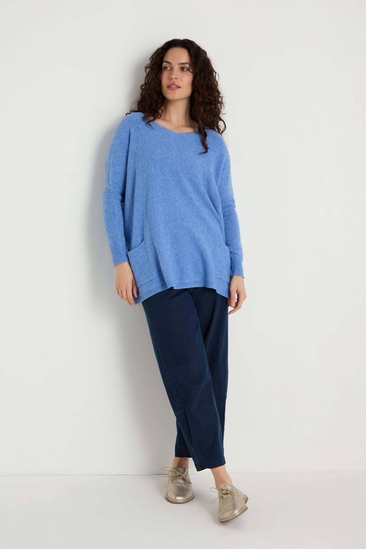 A person stands against a white wall wearing the Two Pocket Oversize Knit in blue, paired with dark blue pants and beige shoes—a perfect example of cozy women's knitwear.